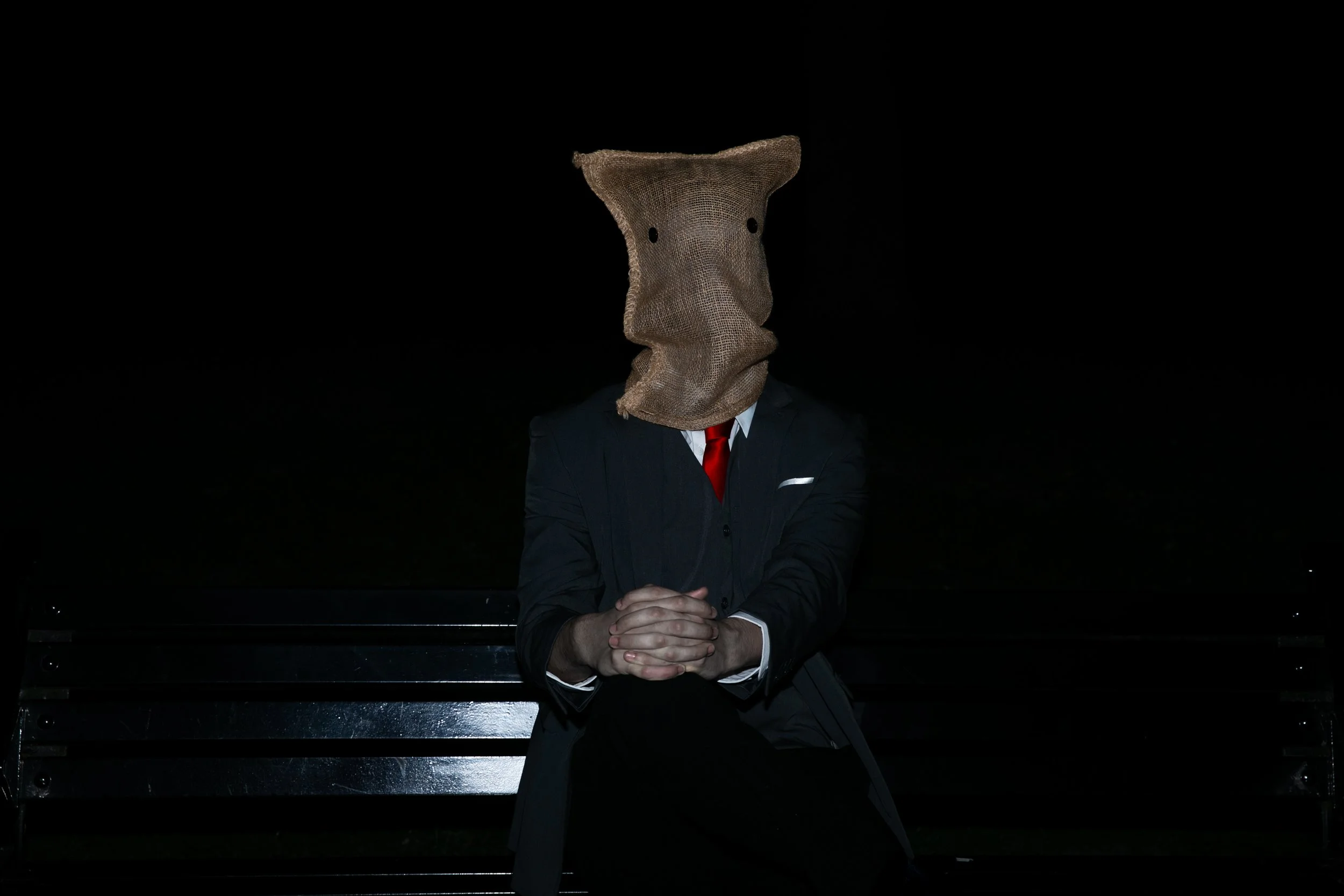 Person in a suit with a red tie, sitting on a bench at night, wearing a burlap sack over their head with two eye holes.