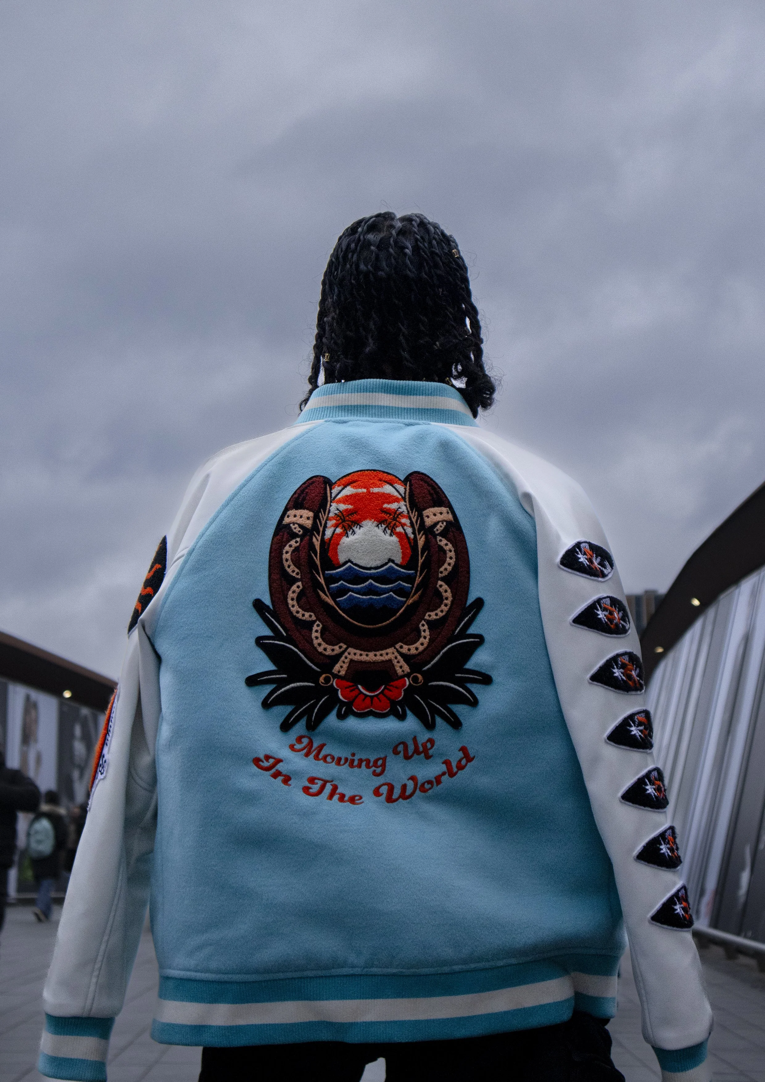 Person with dreadlocks wearing a light blue jacket with a graphic patch on the back that says "Moving Up In The World" and features a design of a sunset over water with palm trees, standing outdoors against a cloudy sky.