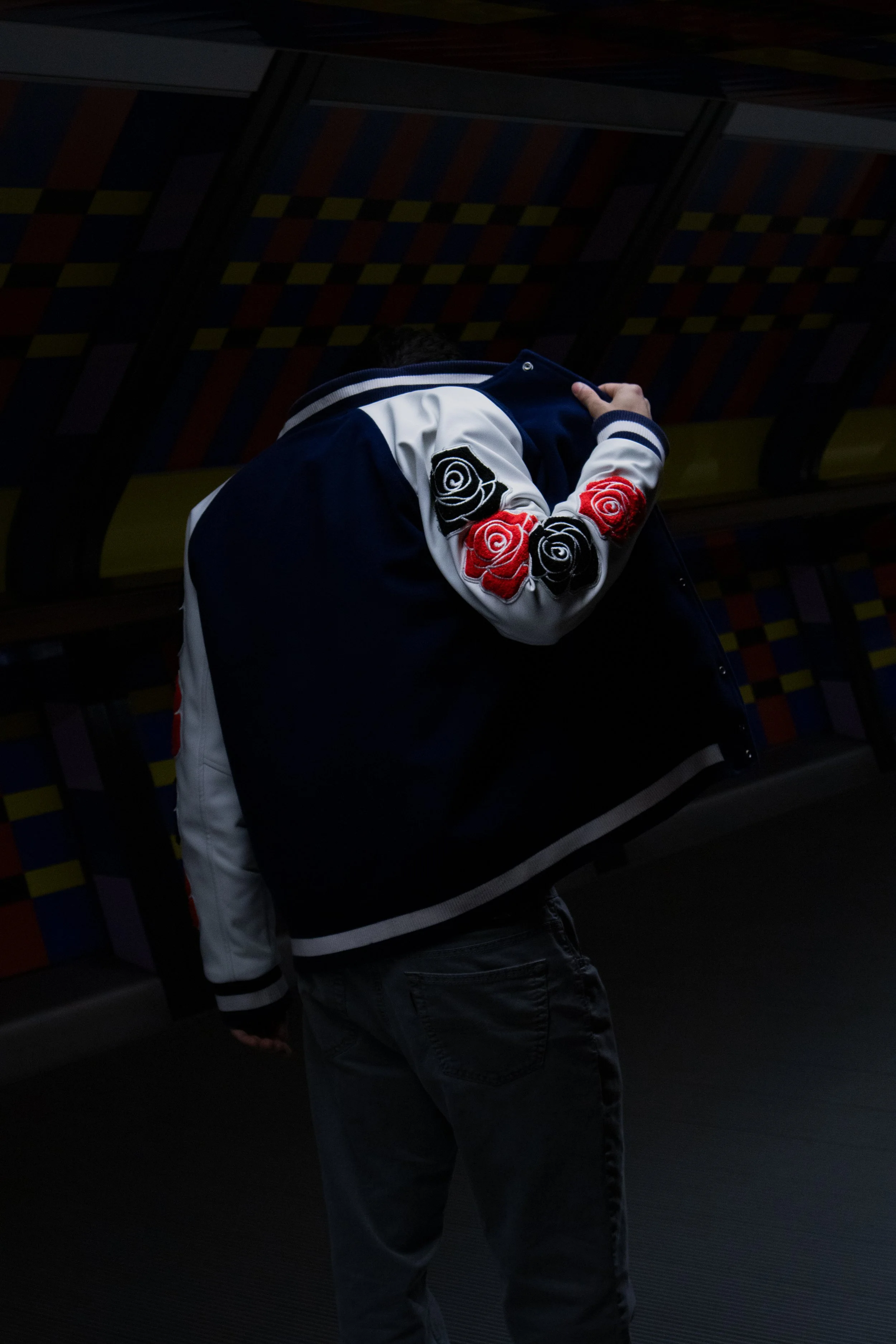 Person wearing a black and white varsity jacket with rose patches on the sleeve, standing in a dimly lit environment with a multi-colored checkered background.