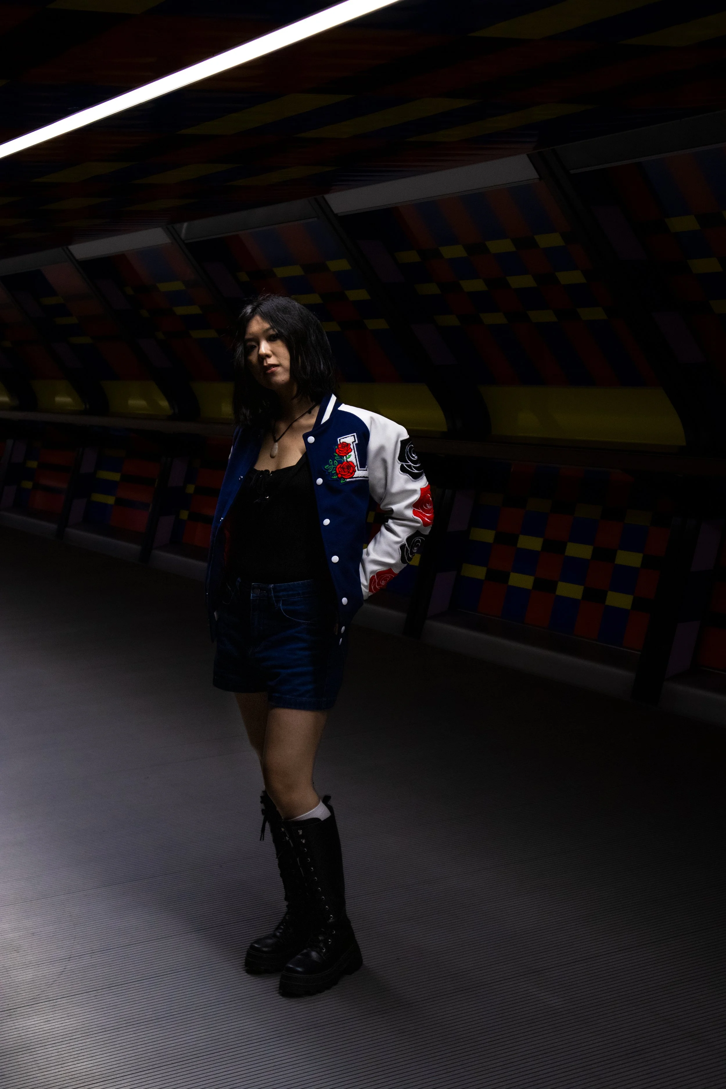 A young woman with black hair, wearing a varsity jacket with floral patches, shorts, and tall black boots, standing in a dimly lit, colorful hallway.