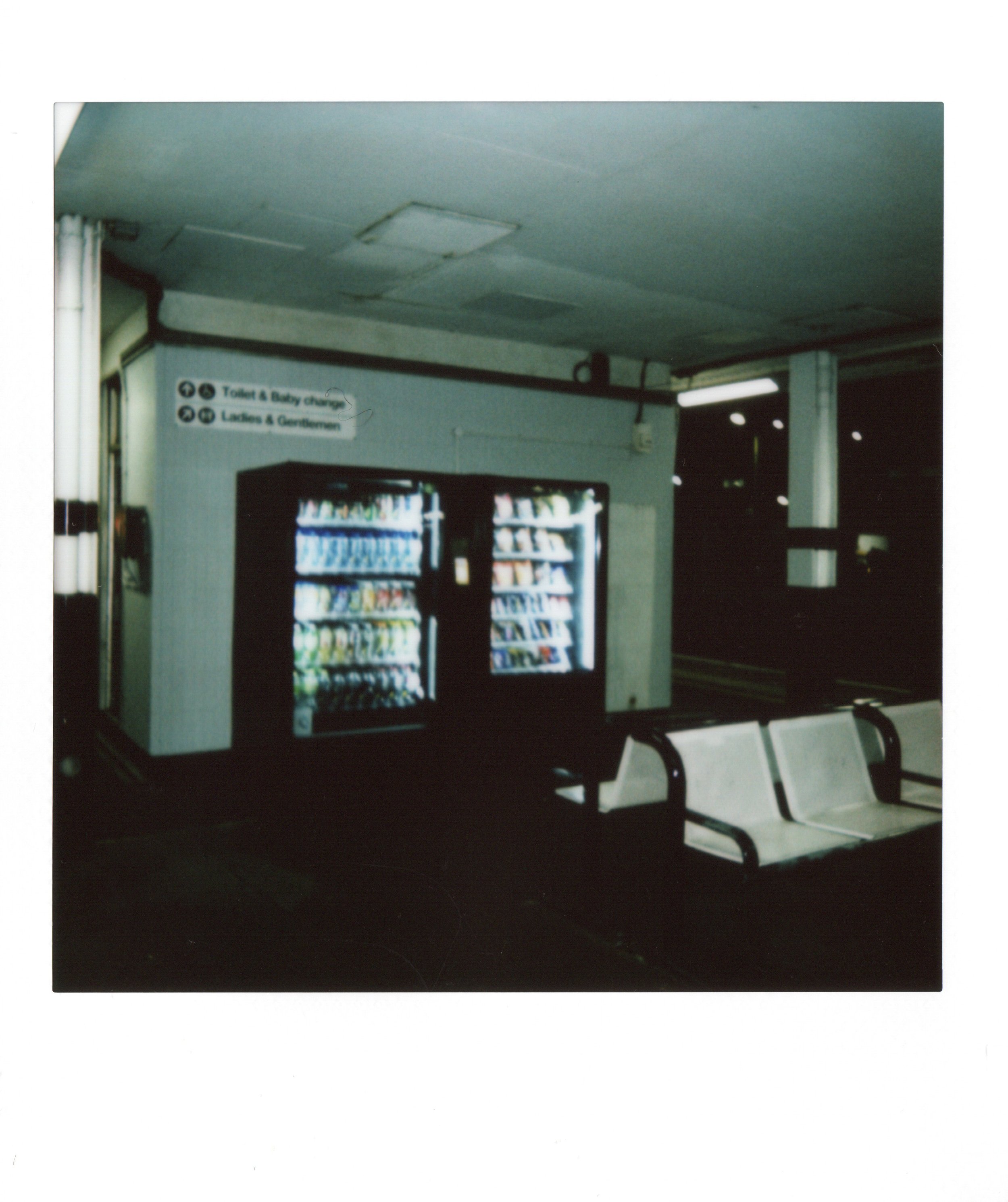 An indoor setting with a vending machine, white bench seats, and signs indicating women's and men's toilets and baby changing, with low light and a slightly blurry image.