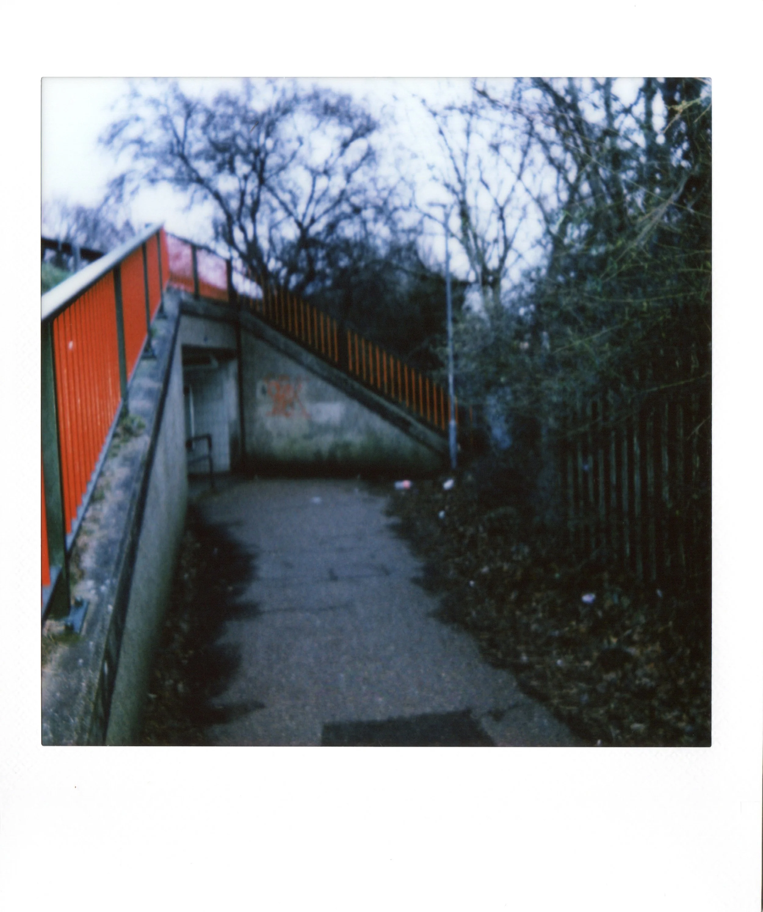 A pathway beneath a bridge with orange and black railing, surrounded by leafless trees and a black metal fence on the right. The scene appears to be overcast or in low light.