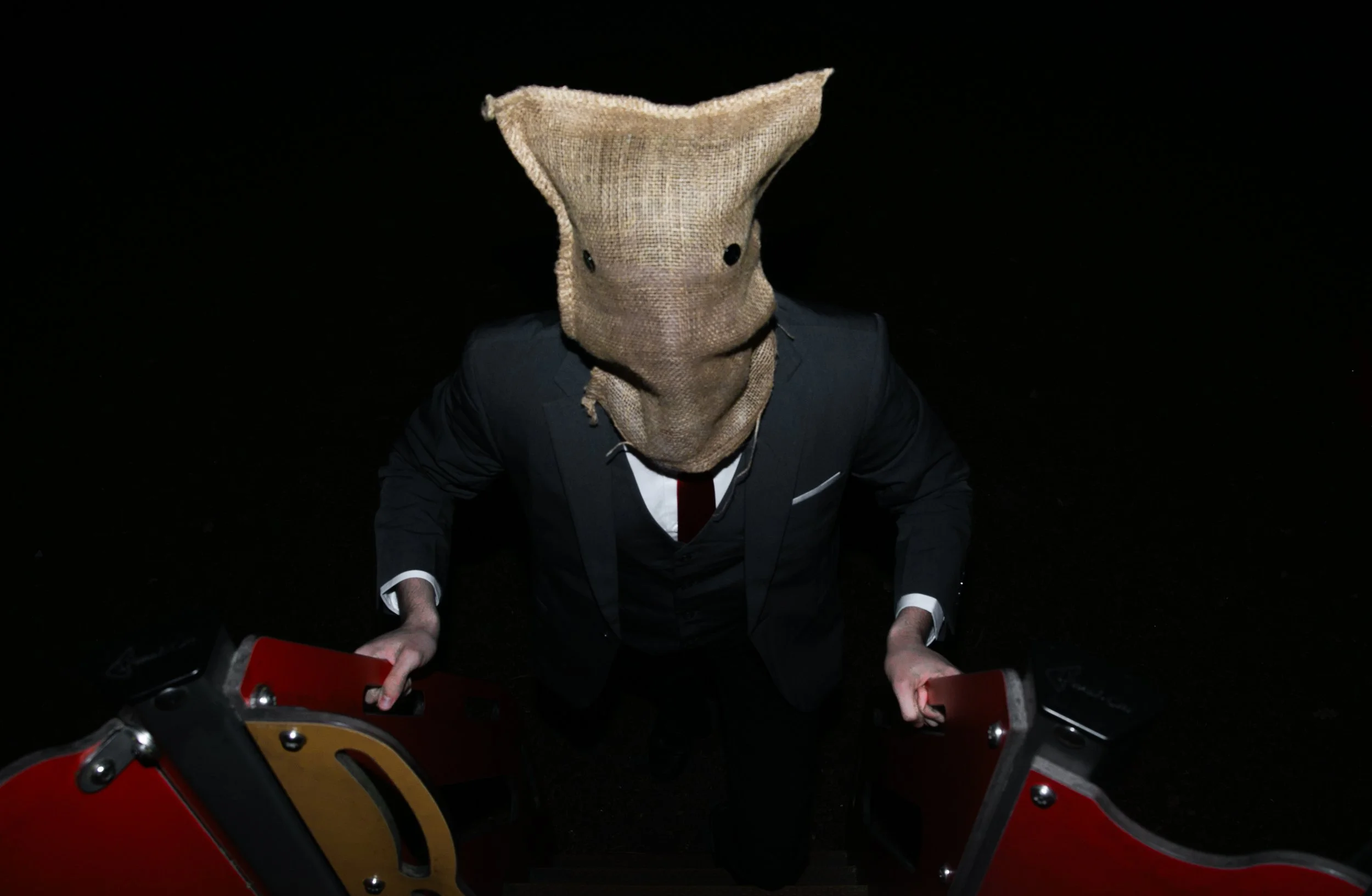 Person wearing a suit and a burlap sack over their head, holding onto the handles of red and black shelves.