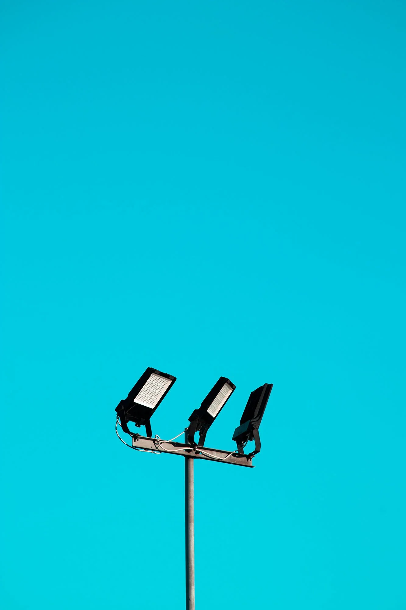 Three outdoor floodlights mounted on a tall pole against a clear blue sky