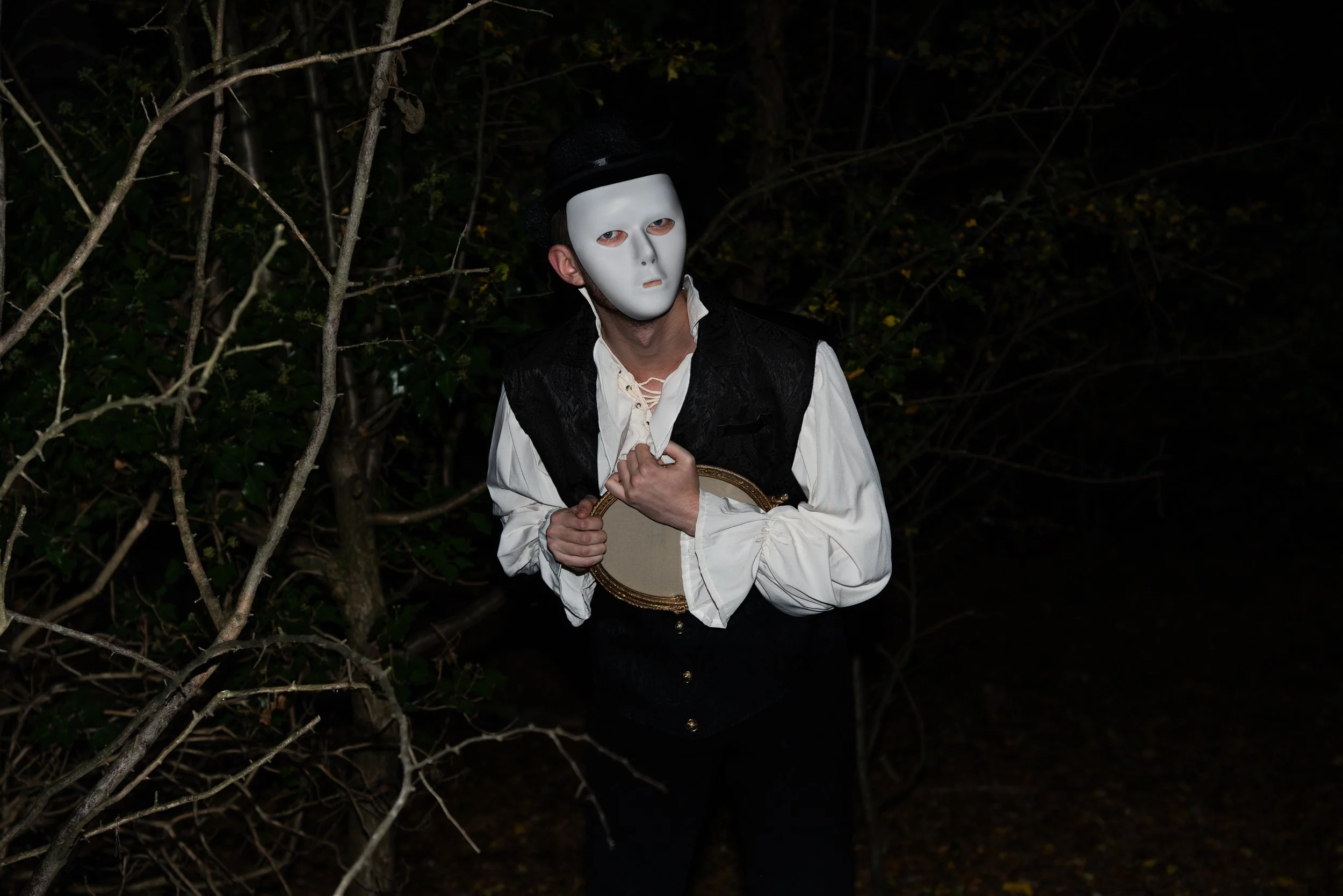 A person wearing a white mask and black hat, dressed in vintage clothing, holding a small mirror, standing in a dark outdoor setting with trees.