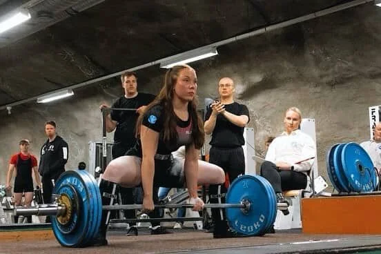 Nuori nainen nostamassa tankoa kuntosalilla, ympärillä muita ihmisiä.