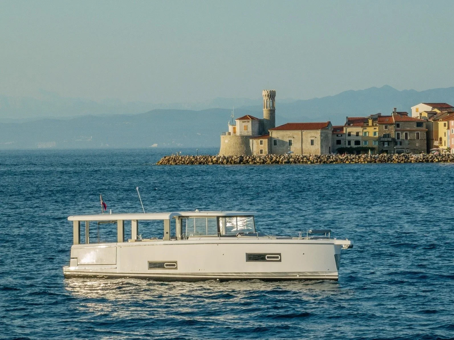 A white motorboat floating on the water with a coastal town and a castle-like structure with a tower on a rocky jetty in the background under a hazy sky.