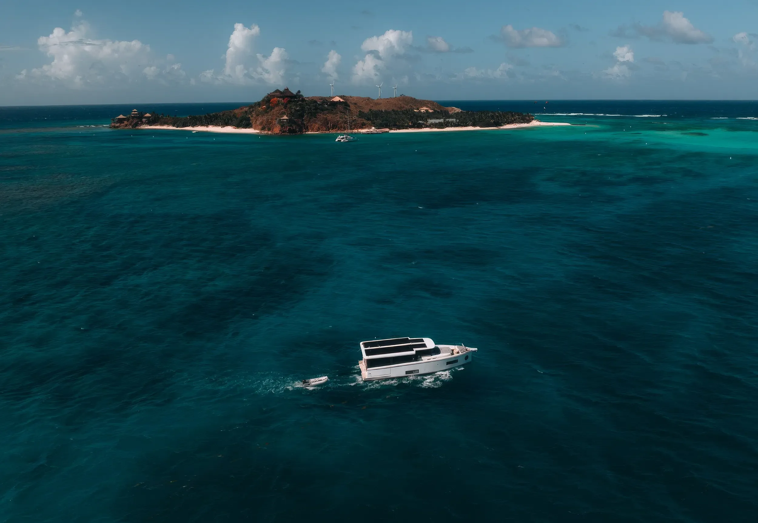 A boat sailing near an island with a coastline, some buildings on a hill, and wind turbines on the hilltop. The water is a shade of turquoise, and the sky is partly cloudy.