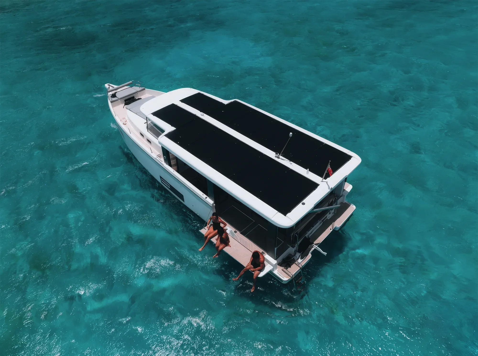 A white yacht with a black roof floating on turquoise water, with three people hanging off the side of the yacht.