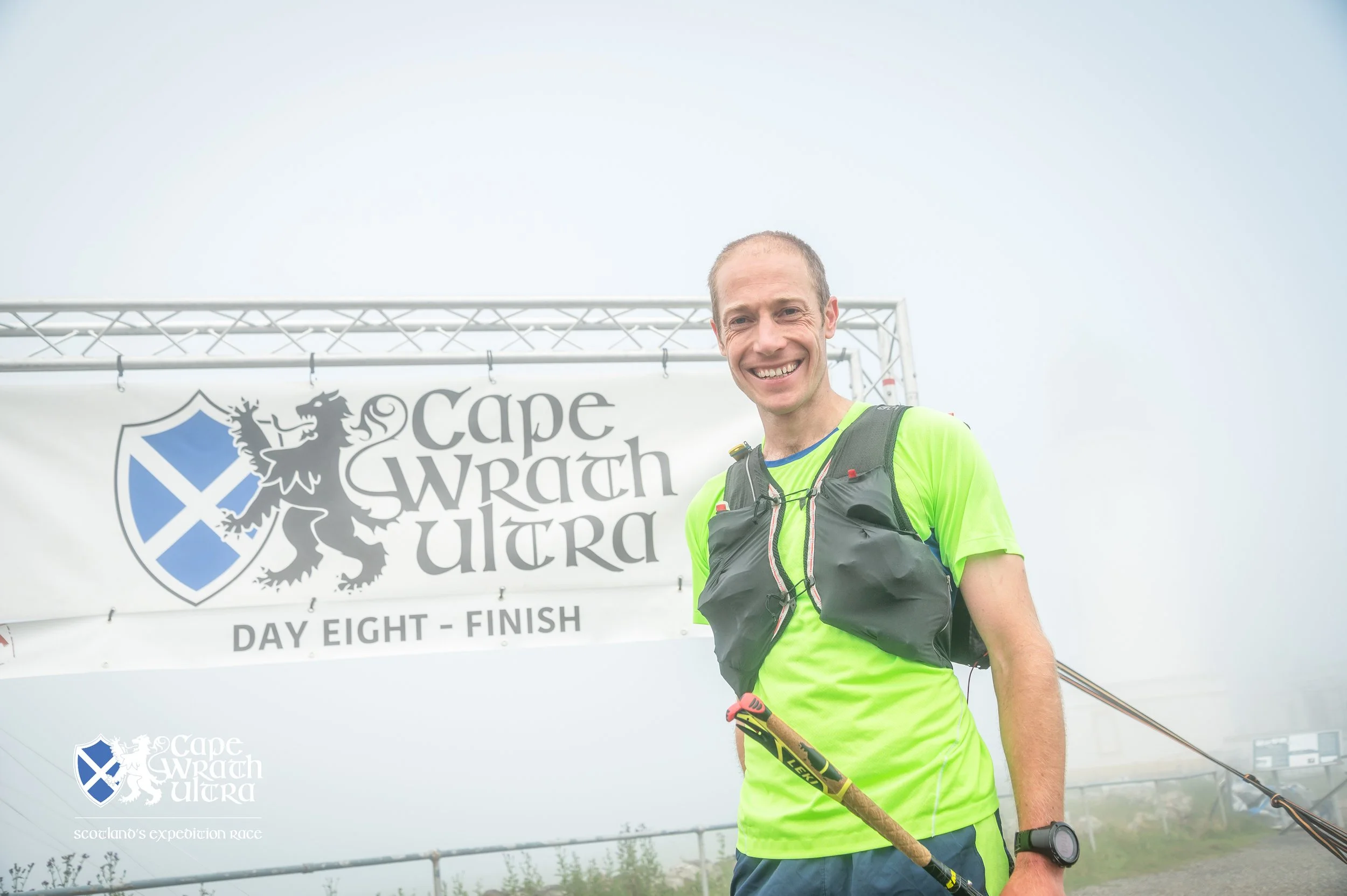 A smiling man in a bright green shirt and black running vest holding trekking poles, standing in front of a banner that reads 'Cape Wrath Ultra,' indicating the finish of day eight of an ultramarathon race.