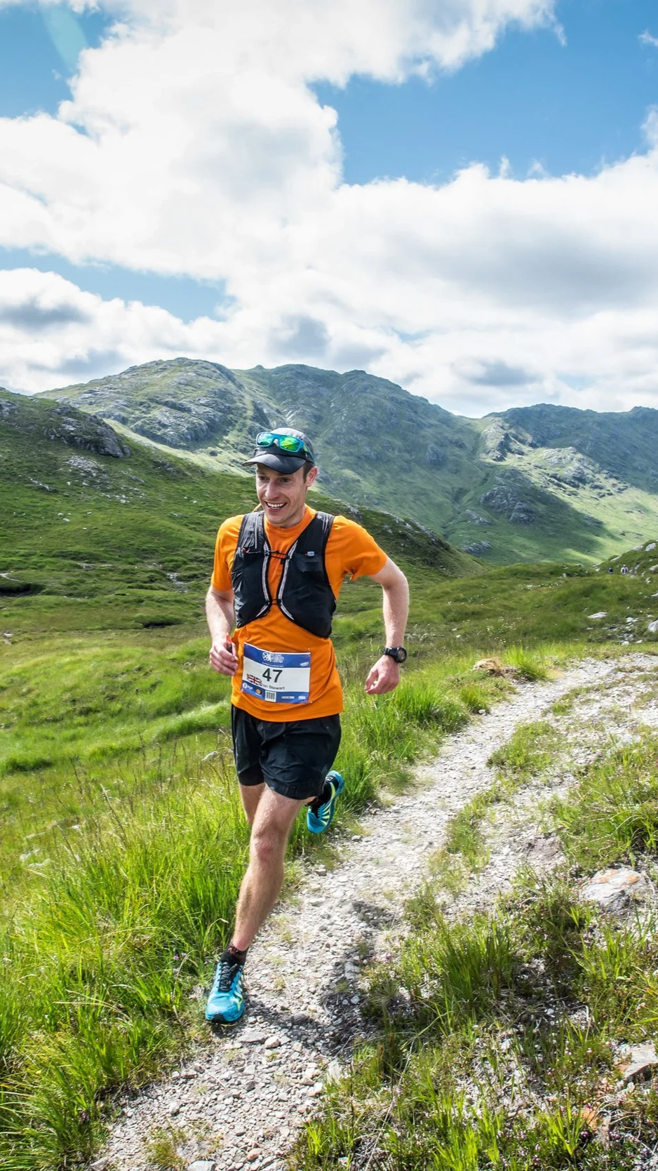 A man running on a narrow trail through a grassy mountainous landscape during a trail race. He is wearing an orange shirt, black shorts, a watch, sunglasses on his head, and a hydration vest. The sky is partly cloudy with blue skies visible.