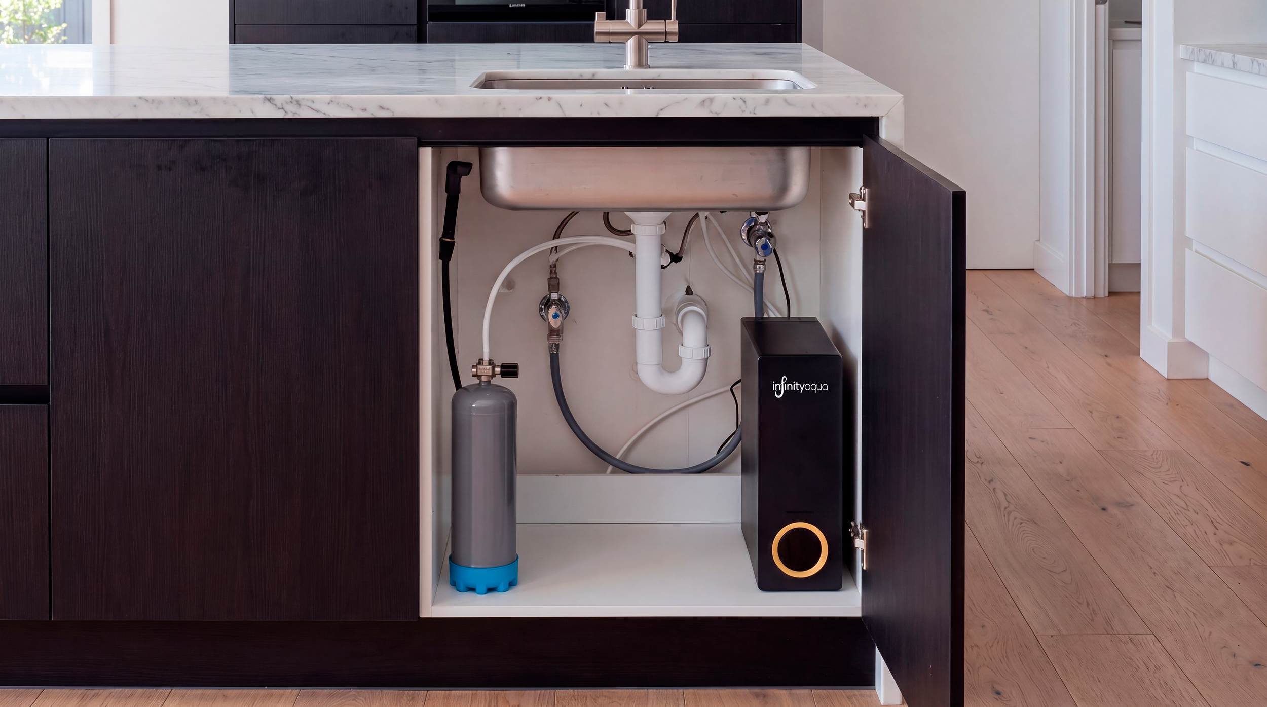 Vista de debajo del fregadero de una cocina con componentes de un sistema de filtración de agua, incluyendo un cilindro de filtración de agua, tuberías de PVC y conexiones, en un gabinete abierto de madera oscura.