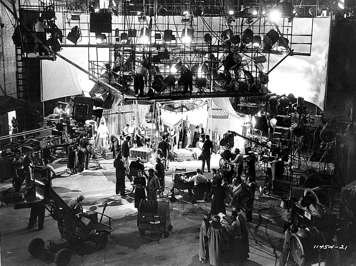Black and white photo of a busy television studio set with crew members, cameras, lights, and other production equipment, as well as actors or performers in costumes.