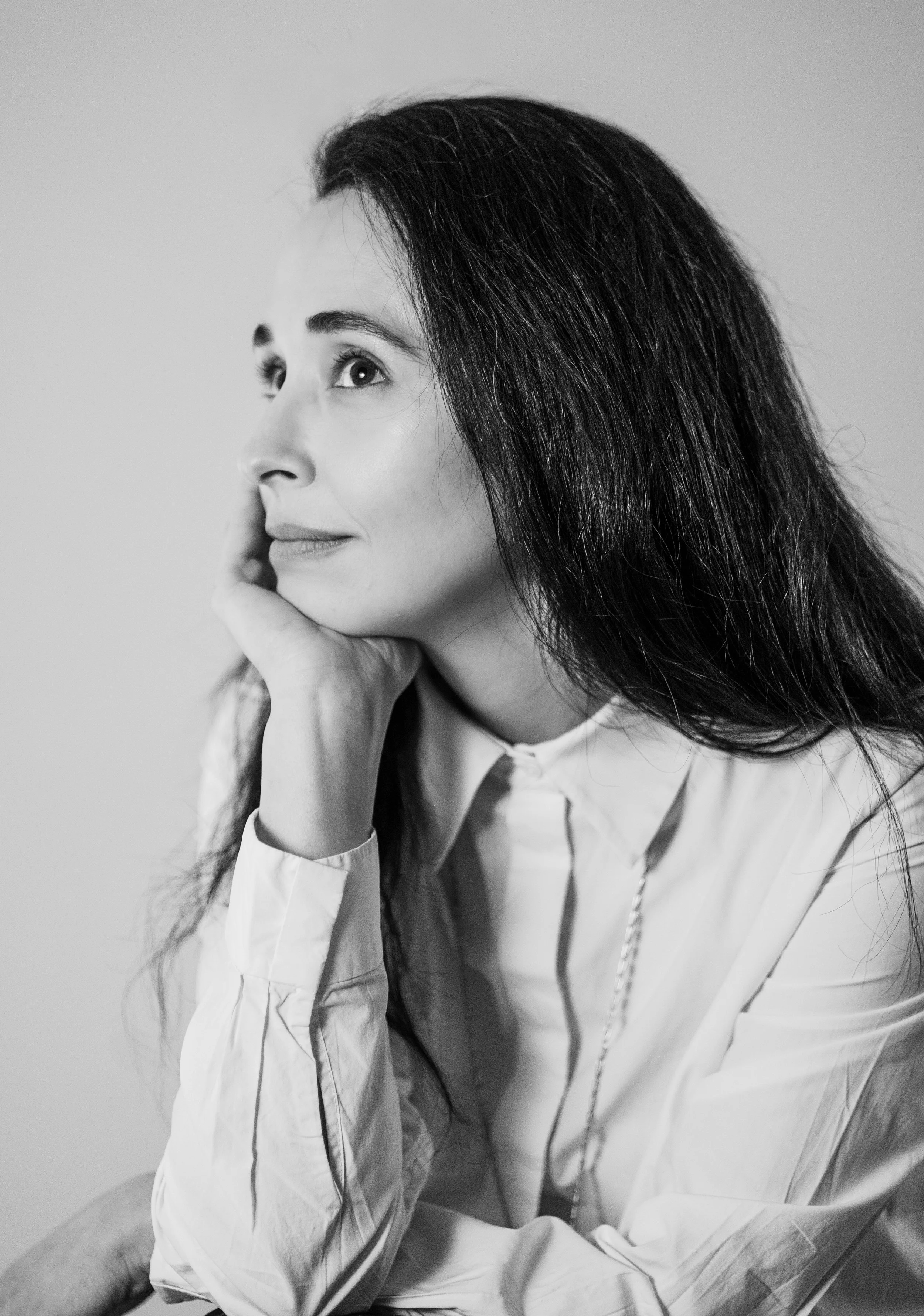 A black-and-white portrait of a woman with long dark hair, resting her chin on her hand and looking upward.
