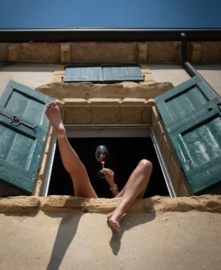 Person relaxing in a window opening with turquoise shutters, holding a glass of red wine.