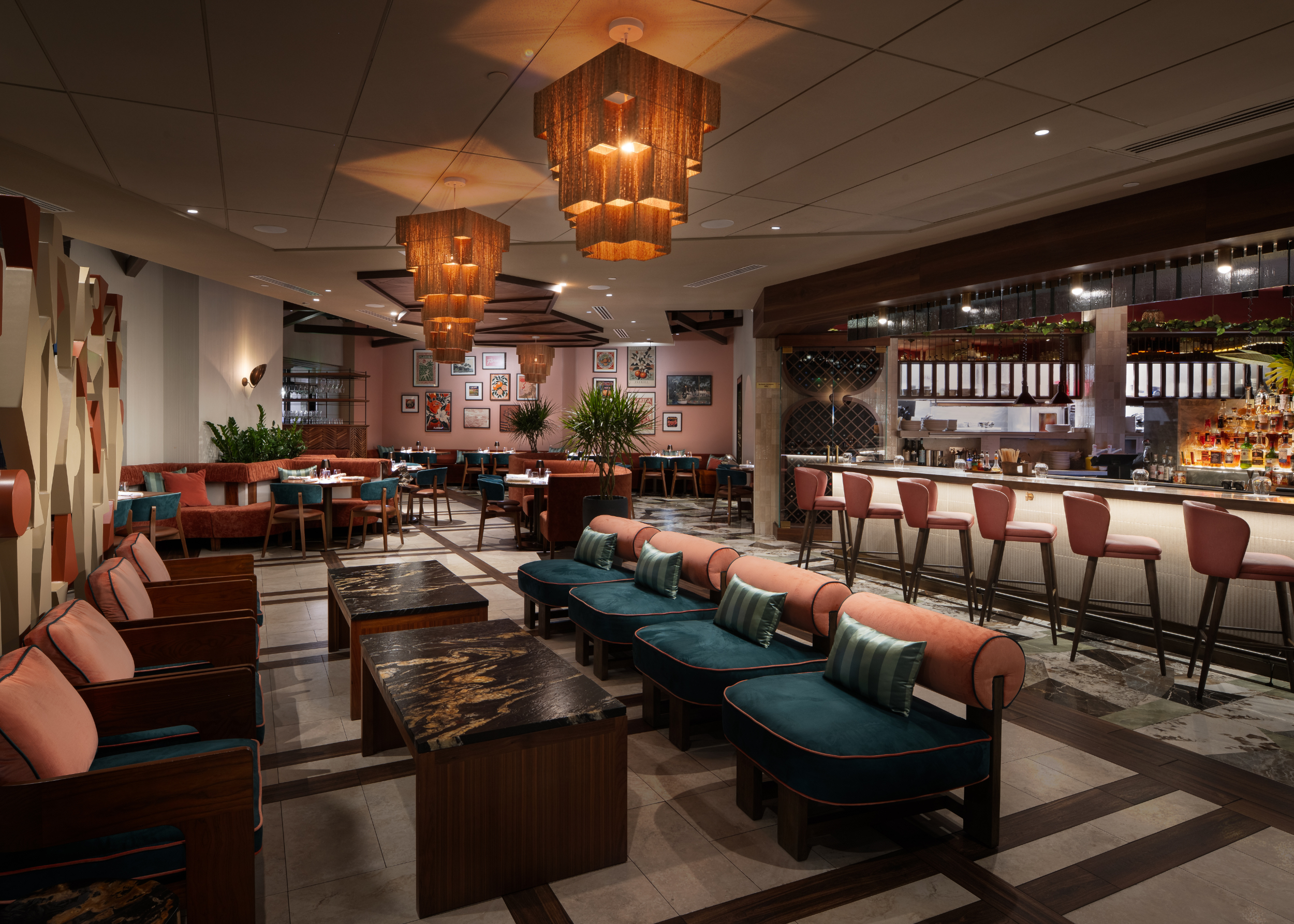 Interior of a modern restaurant with hanging brown pendant lights, a bar area, and framed artwork on the pink wall.
