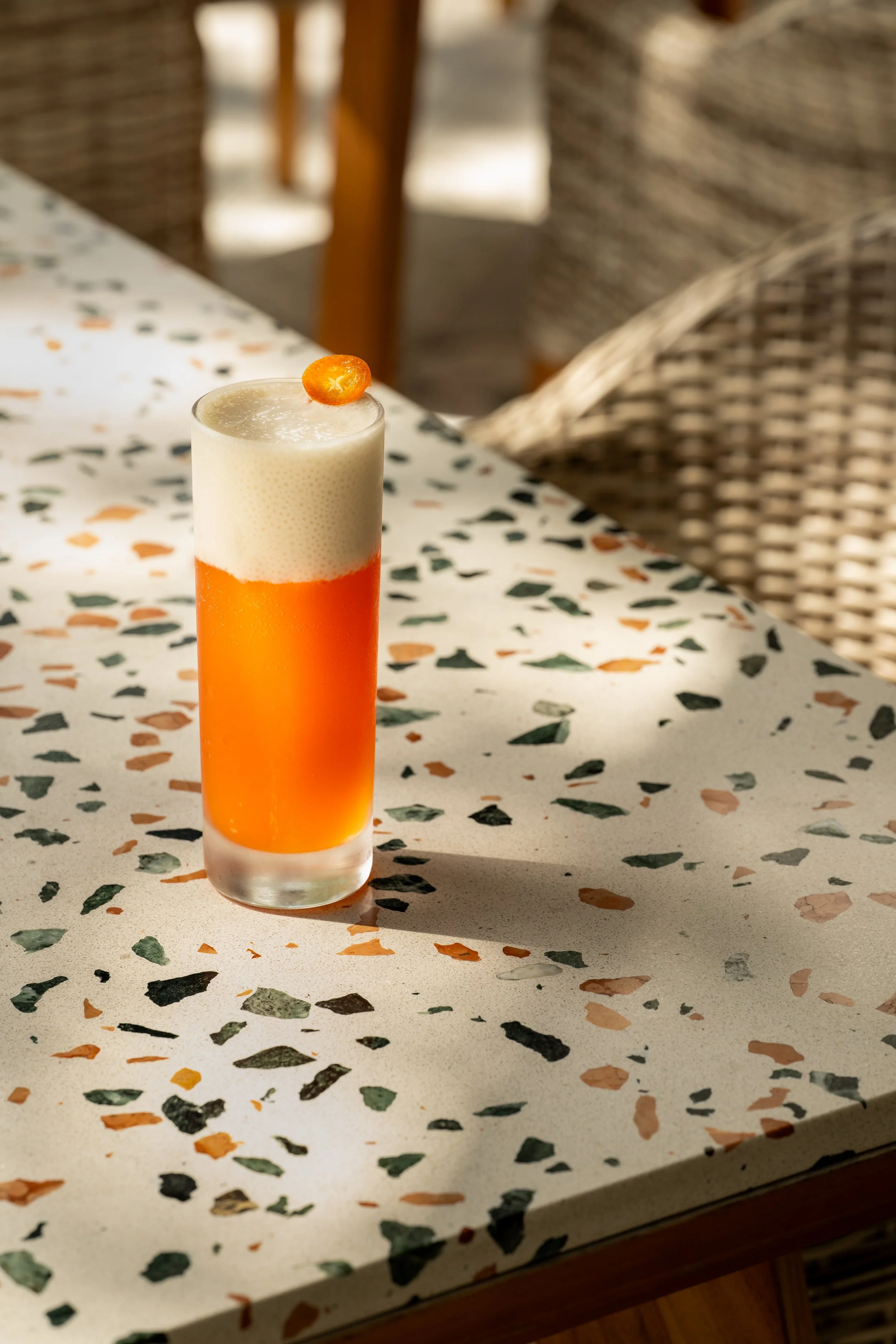 Sunshine State - One of The Grove Bar + Kitchen Signature Cocktails - A tall glass of orange and cream-colored layered beverage with a small orange slice on top, placed on a terrazzo table with scattered green, orange, black, and white flecks.