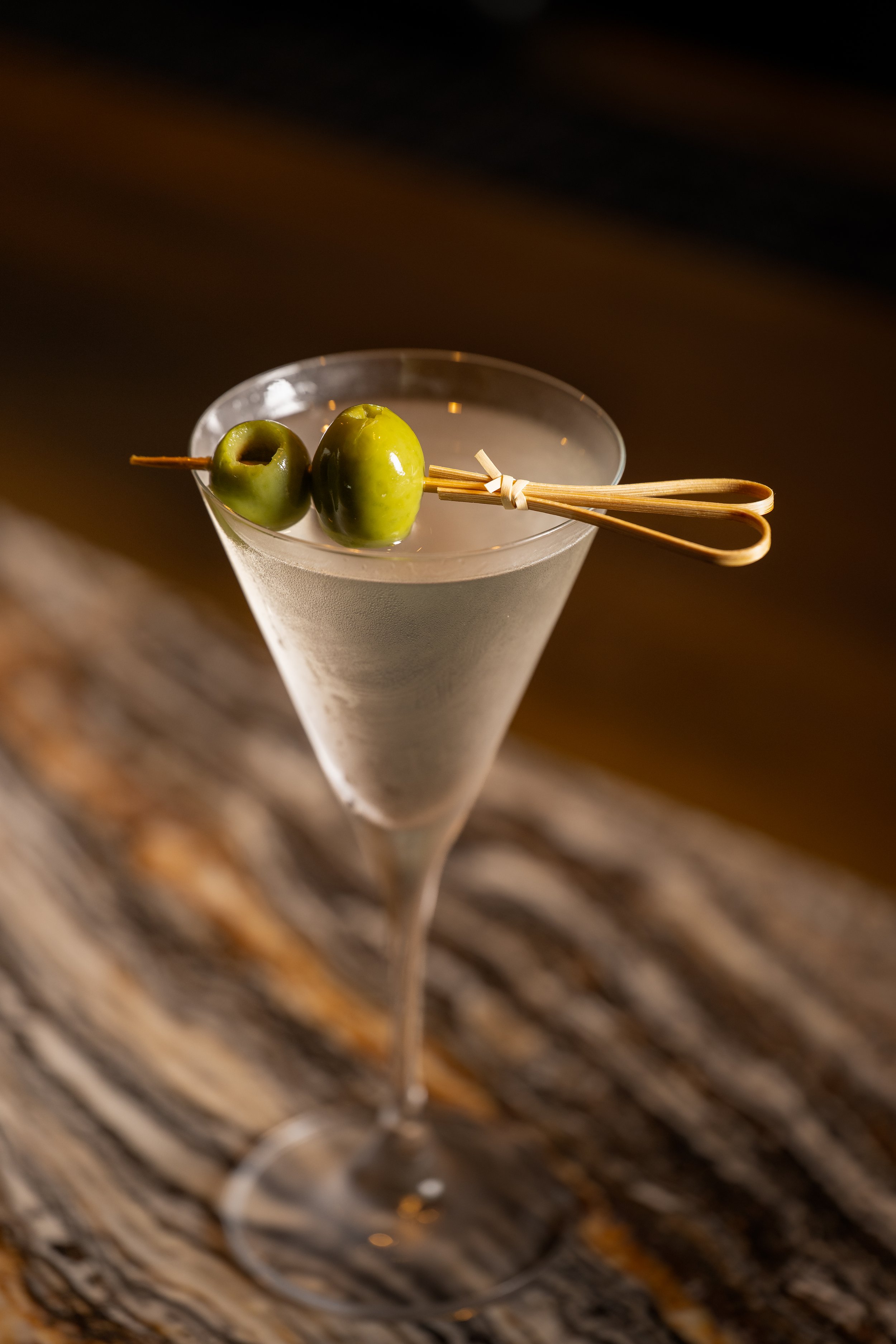 Close-up of a clear martini glass with two green olives on a wooden skewering pick, resting on a wooden surface.