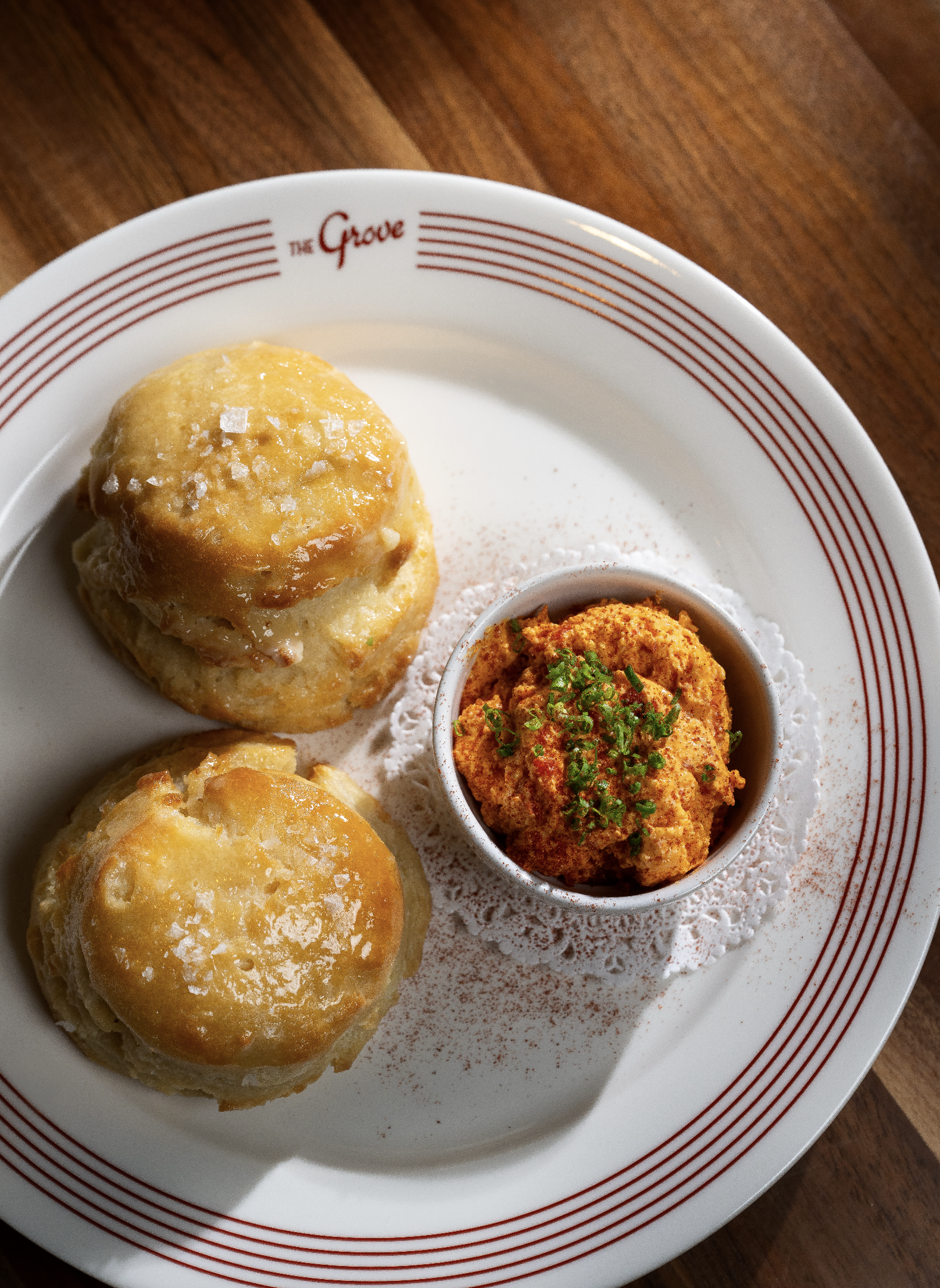 A white plate with red stripes around the rim and the words 'The Grove' printed on it, holding two honey biscuits with coarse salt on top and a small bowl of pimento cheese spread, garnished with chopped herbs.