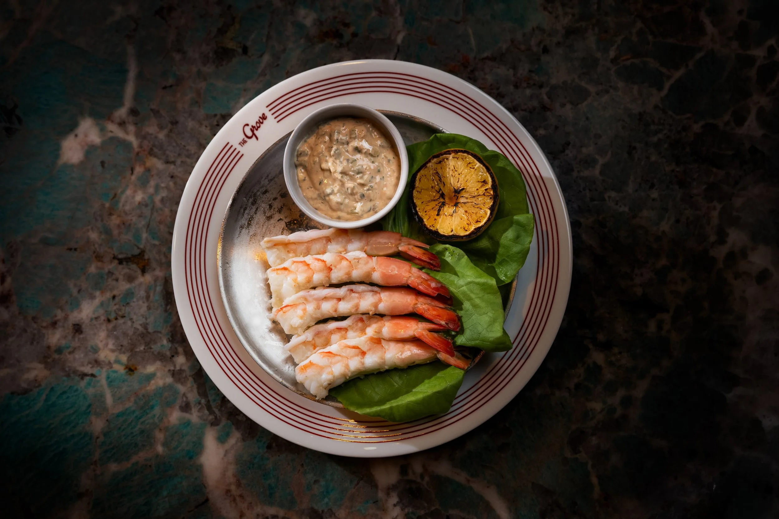Plate with cooked shrimp, a lemon half, and a small bowl of dipping sauce on a dark, patterned surface.