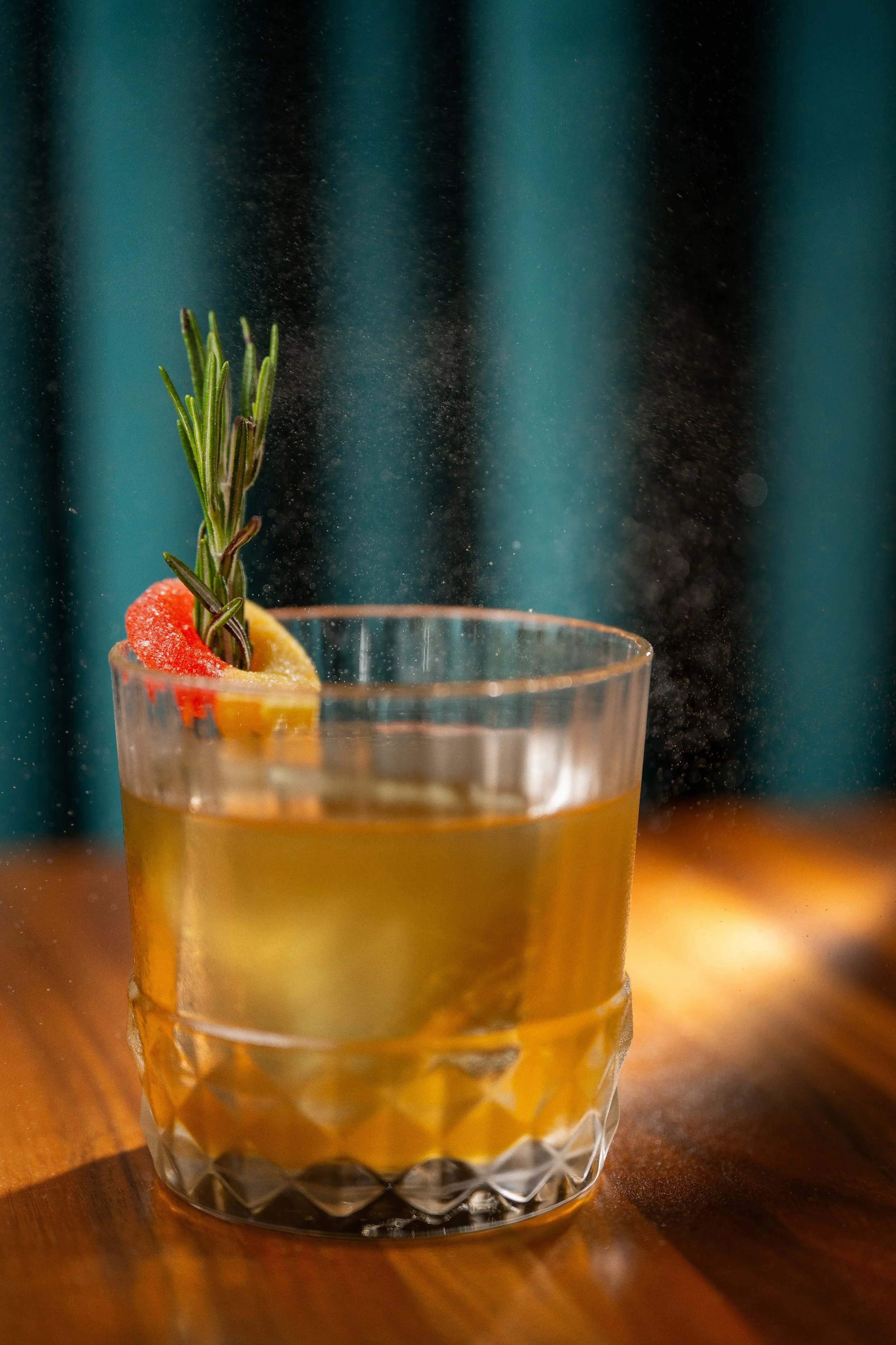 A clear glass with a textured pattern filled with a yellow liquid, garnished with a sprig of rosemary and slices of red and yellow citrus fruits.
