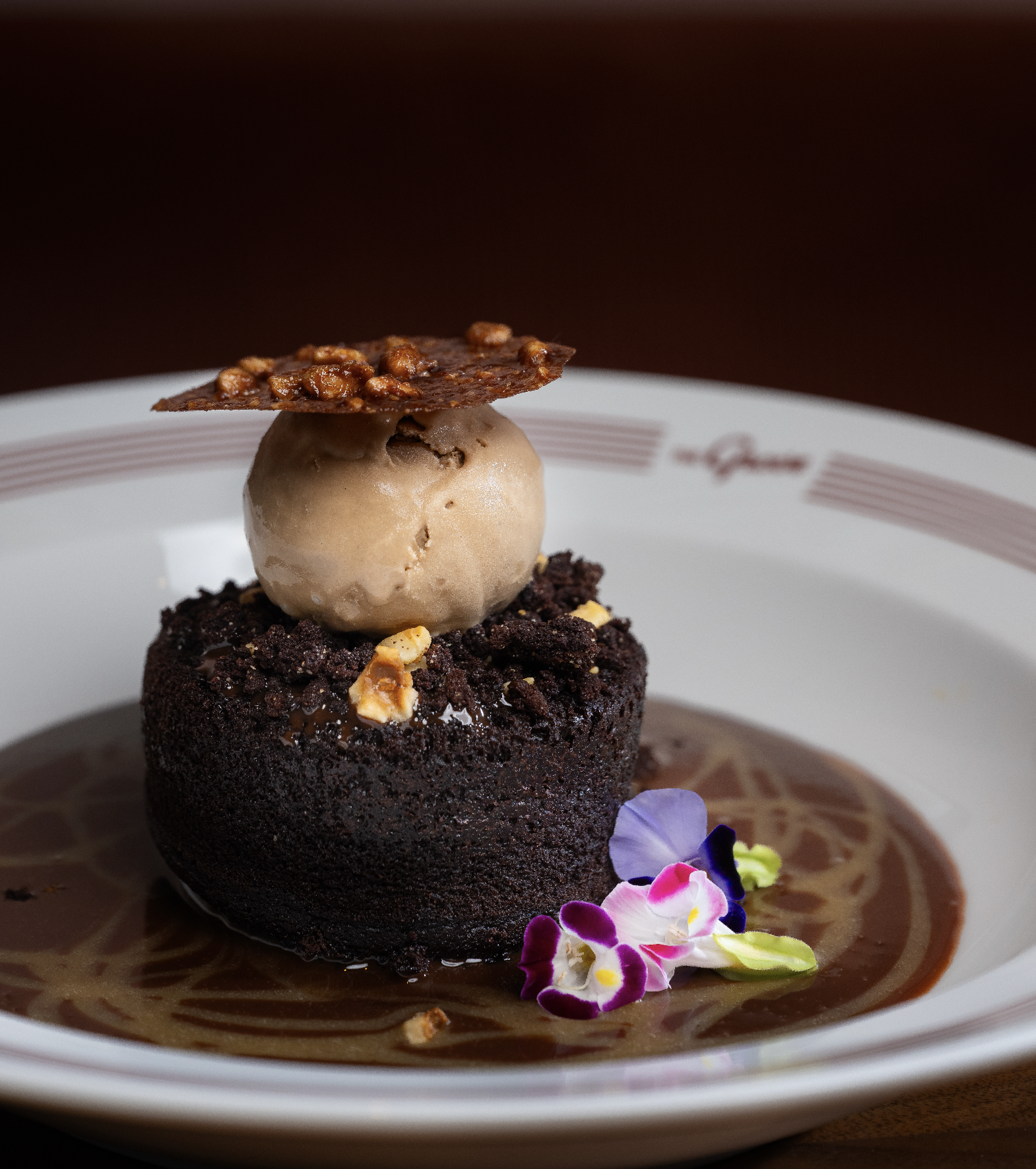 An elaborately plated dessert featuring a chocolate cake, a scoop of ice cream topped with a nut brittle, and edible flowers surrounded by a dark sauce on a white plate.