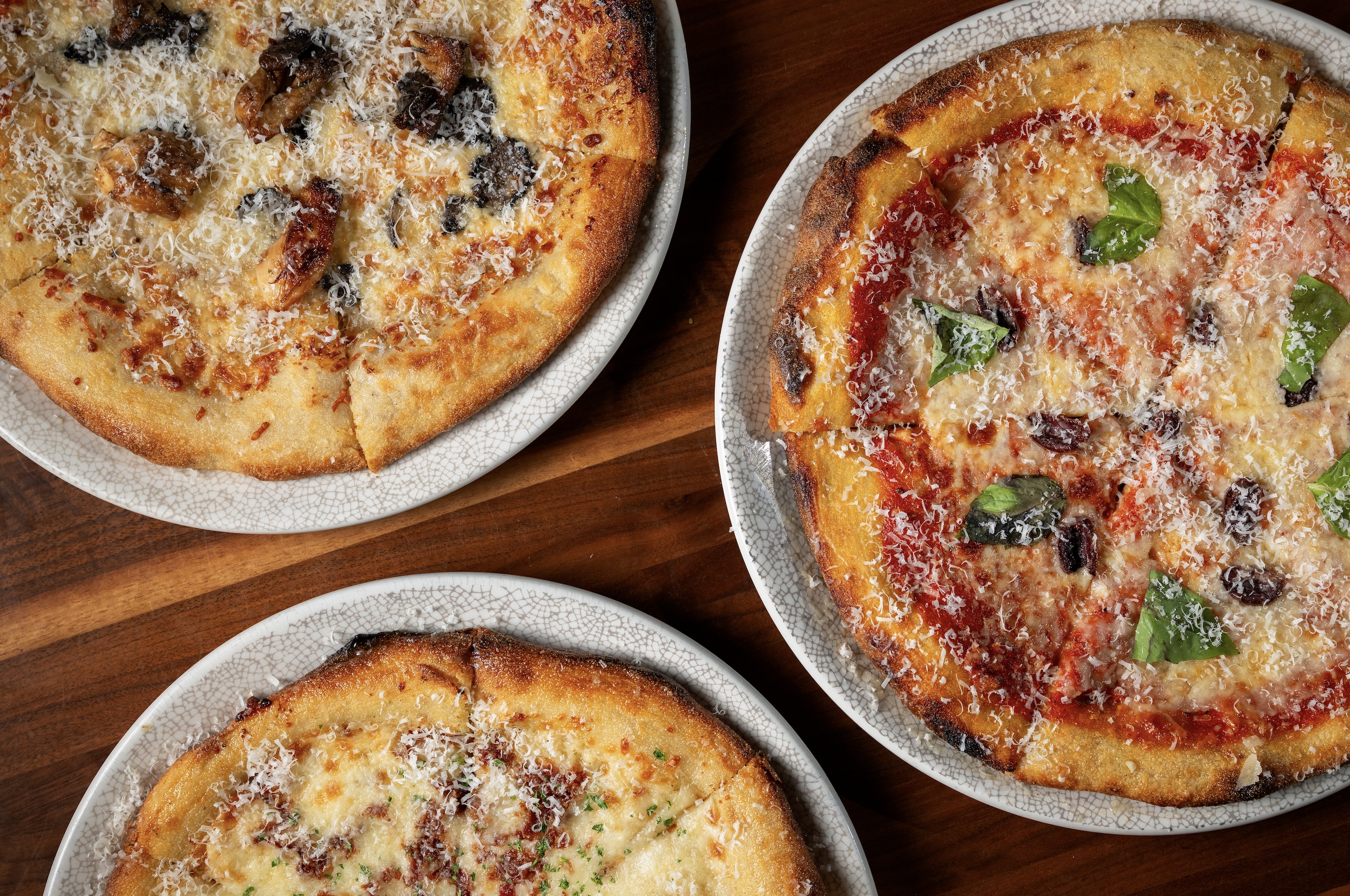 Four plates of different types of pizza on a wooden surface. One pizza has mushrooms and cheese, another has basil leaves, cheese, and tomato sauce, and two others are partially visible with cheese and toppings.
