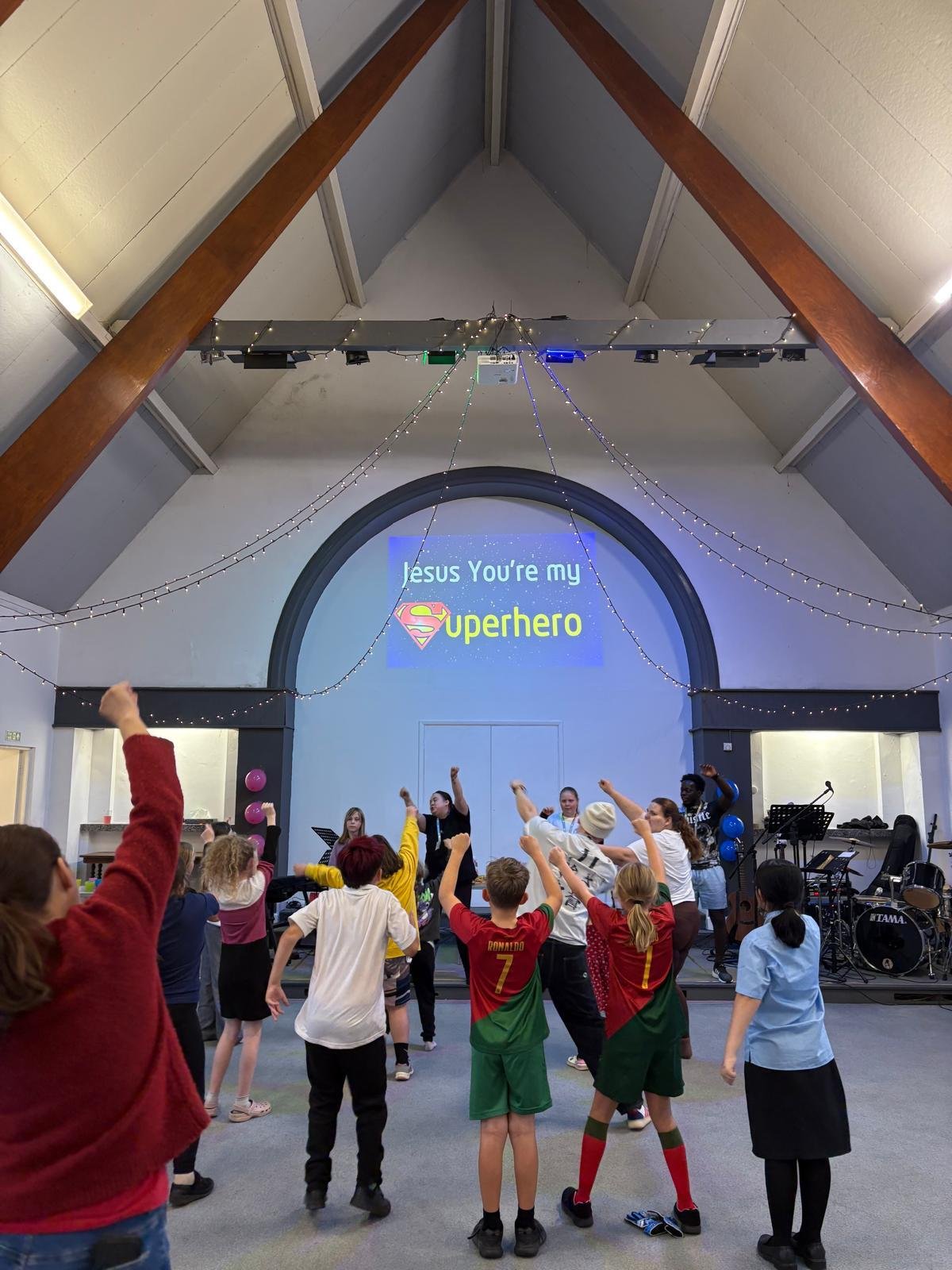 A group of children and adults participating in a church event or celebration, with some raising their fists in the air. The background features a large screen with the message 'Jesus You're my superhero' and a Superman logo, decorated with string lights and balloons.