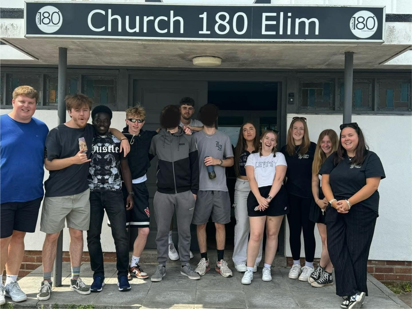Group of eleven young people standing outside a building with a sign that reads 'Church 180 Elim'.