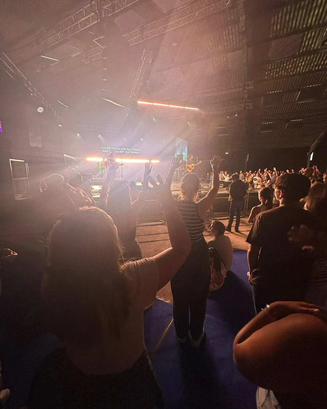 Audience at a live music concert with some people raising their hands, stage with performers, colorful lighting, and a screen displaying song lyrics in the background.