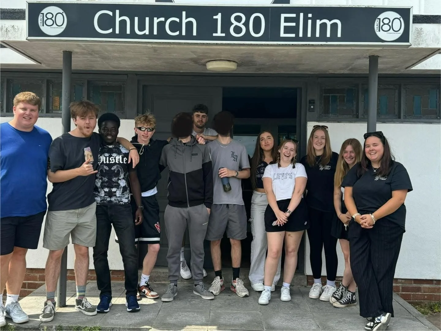 A group of eleven young people standing in front of a church with a sign that reads "Church 180 Elim." They are smiling and appear to be enjoying each other's company. The group is casually dressed, with some holding drinks, and standing on a small porch or entrance area.