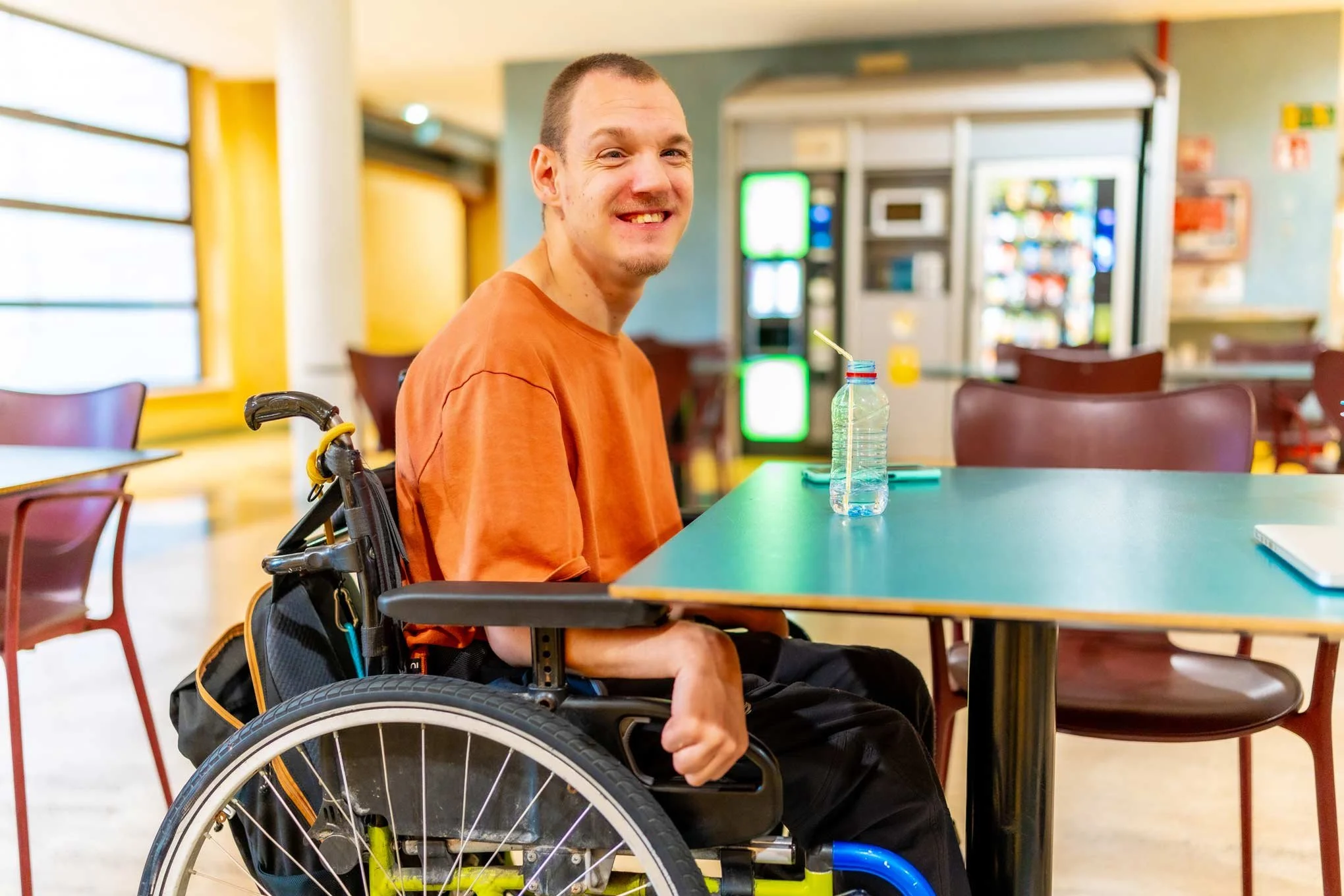 NADSN | National Association of Disabled Staff Networks | A Smiling Man Sitting in a Wheelchair at a Table in a Bright, Modern Cafeteria