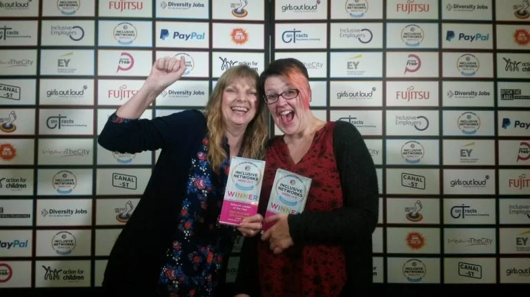 NADSN | National Association of Disabled Staff Networks | Cath Rose and Gill Cockburn smiling and holding awards in front of a backdrop featuring logos for companies like Fujitsu, PayPal and EY