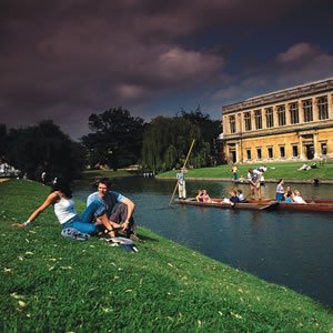 NADSN | National Association of Disabled Staff Networks | A Couple Relaxed on a Grassy River Back With People Punting Past a Historic Building Under a Dramatic Sky