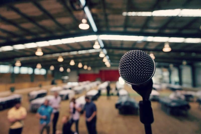 NADSN | National Association of Disabled Staff Networks | A Microphone on a Stand in a Large, Dimly Lit Warehouse With People Blurred in the Background.