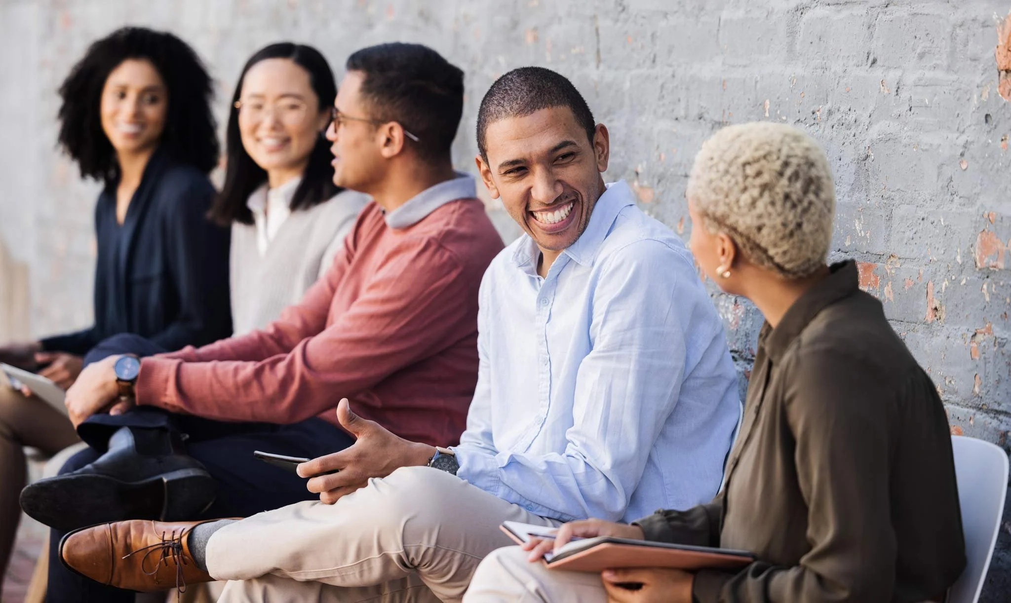 NADSN | National Association of Disabled Staff Networks | A group of diverse young professionals sitting against a grey brick wall, smiling and talking