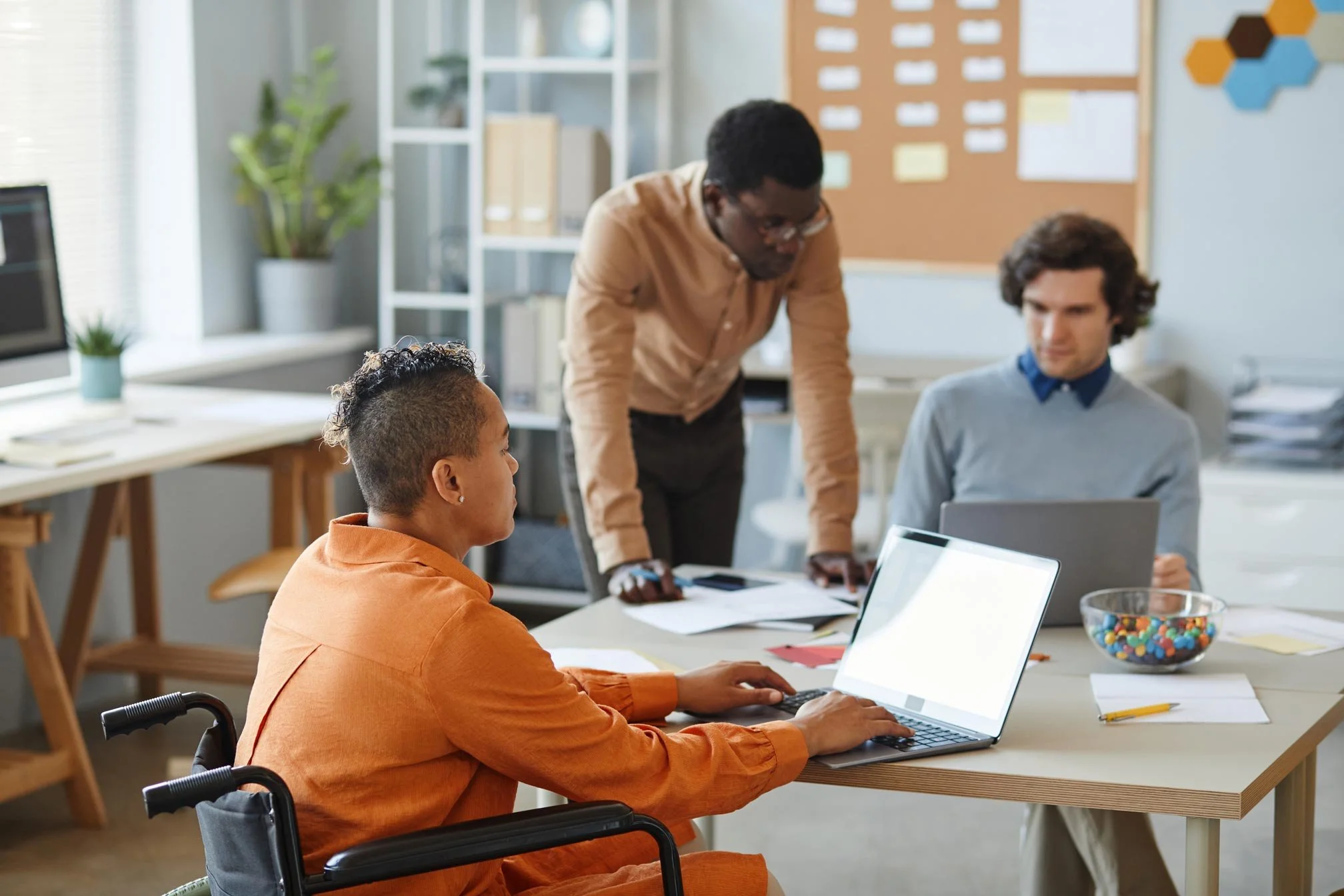 NADSN | National Association of Disabled Staff Networks | Three Colleagues, Including One Person in a Wheelchair, Collaborating on Projects in an Office with Laptops and a Bulletin Board.