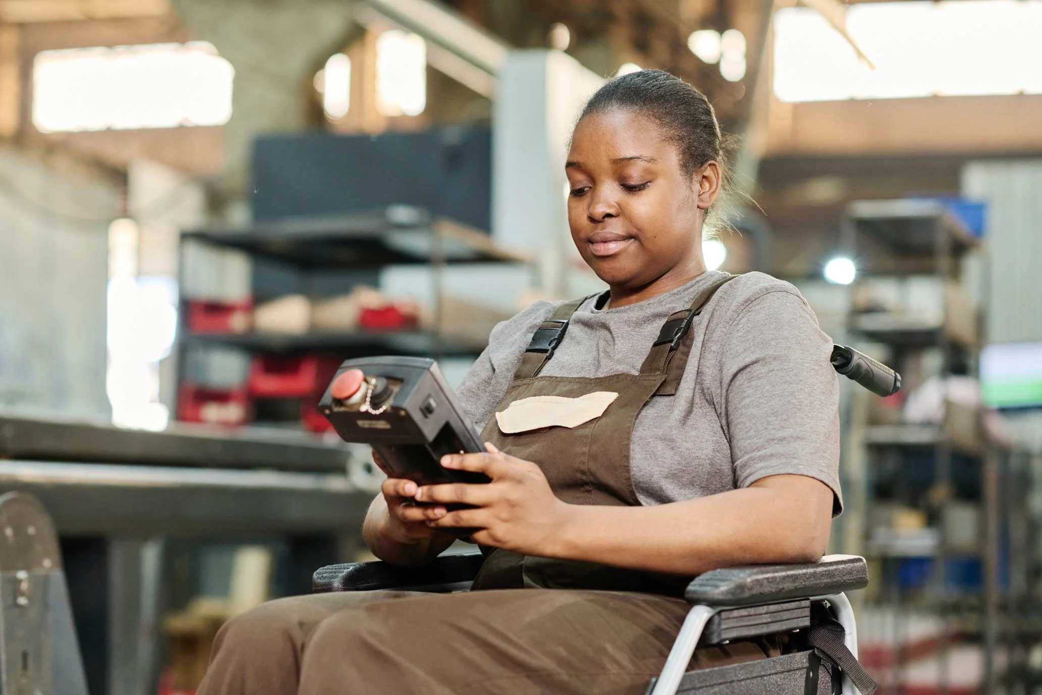 NADSN | National Association of Disabled Staff Networks | A Person in a Wheelchair Wearing Brown Overalls and a Grey T-Shirt Operating a Handheld Control Device
