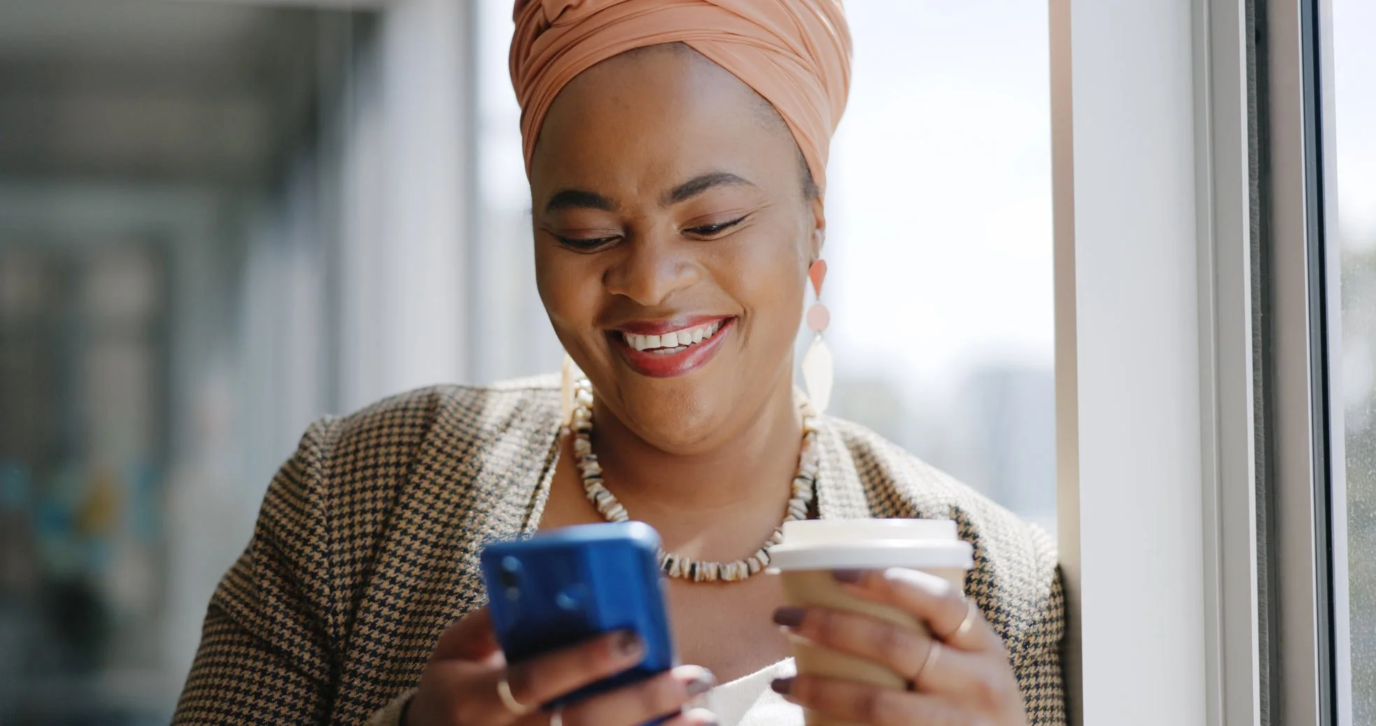 Happy black woman smiling whilst holding a coffee and looking at her phone | NADSN
