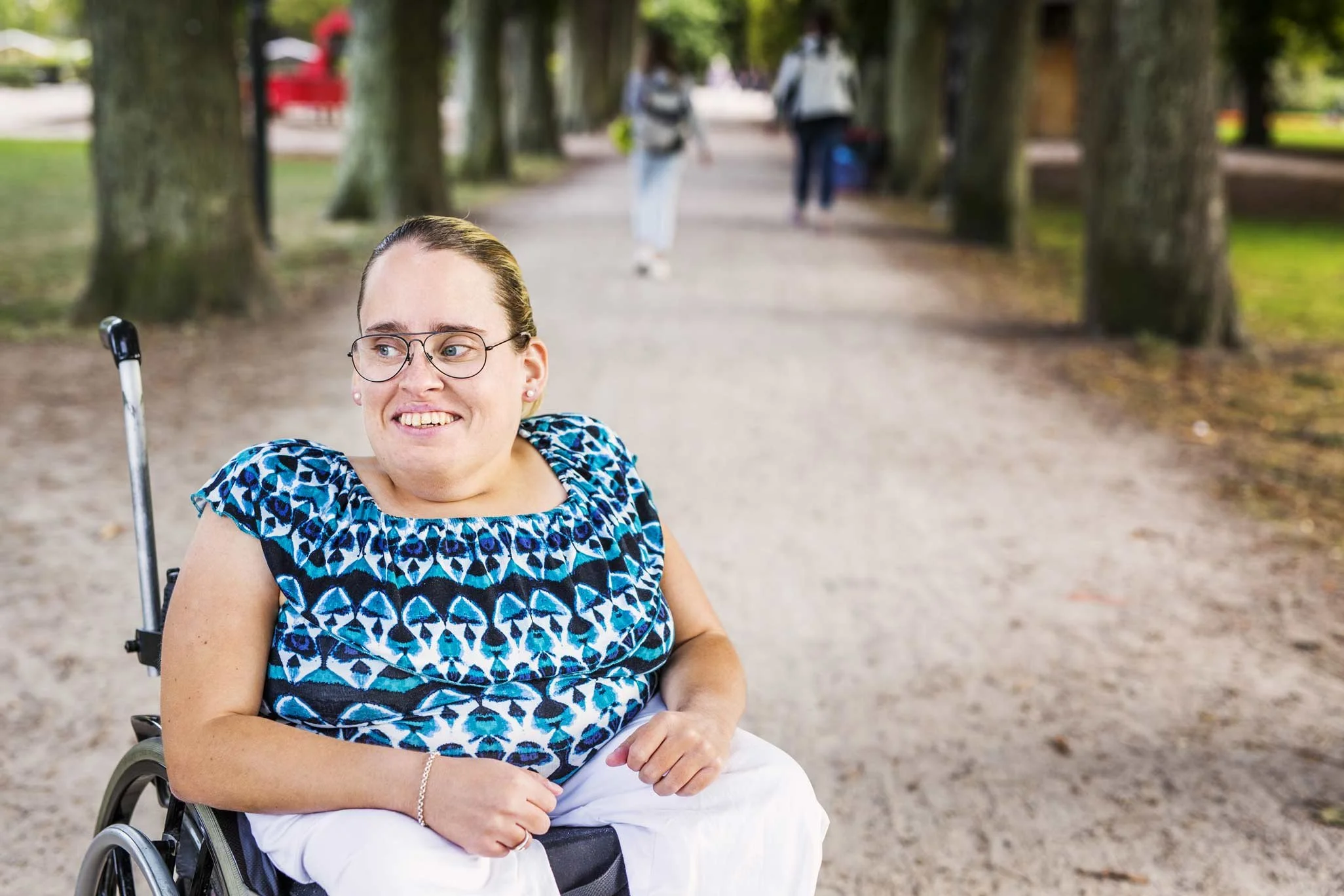 NADSN | National Association of Disabled Staff Networks |  A Woman With Blonde Hair and Glasses Sitting in a Wheelchair Outdoors on a Path Lined With Trees, Wearing a Blue and White Patterned Blouse.