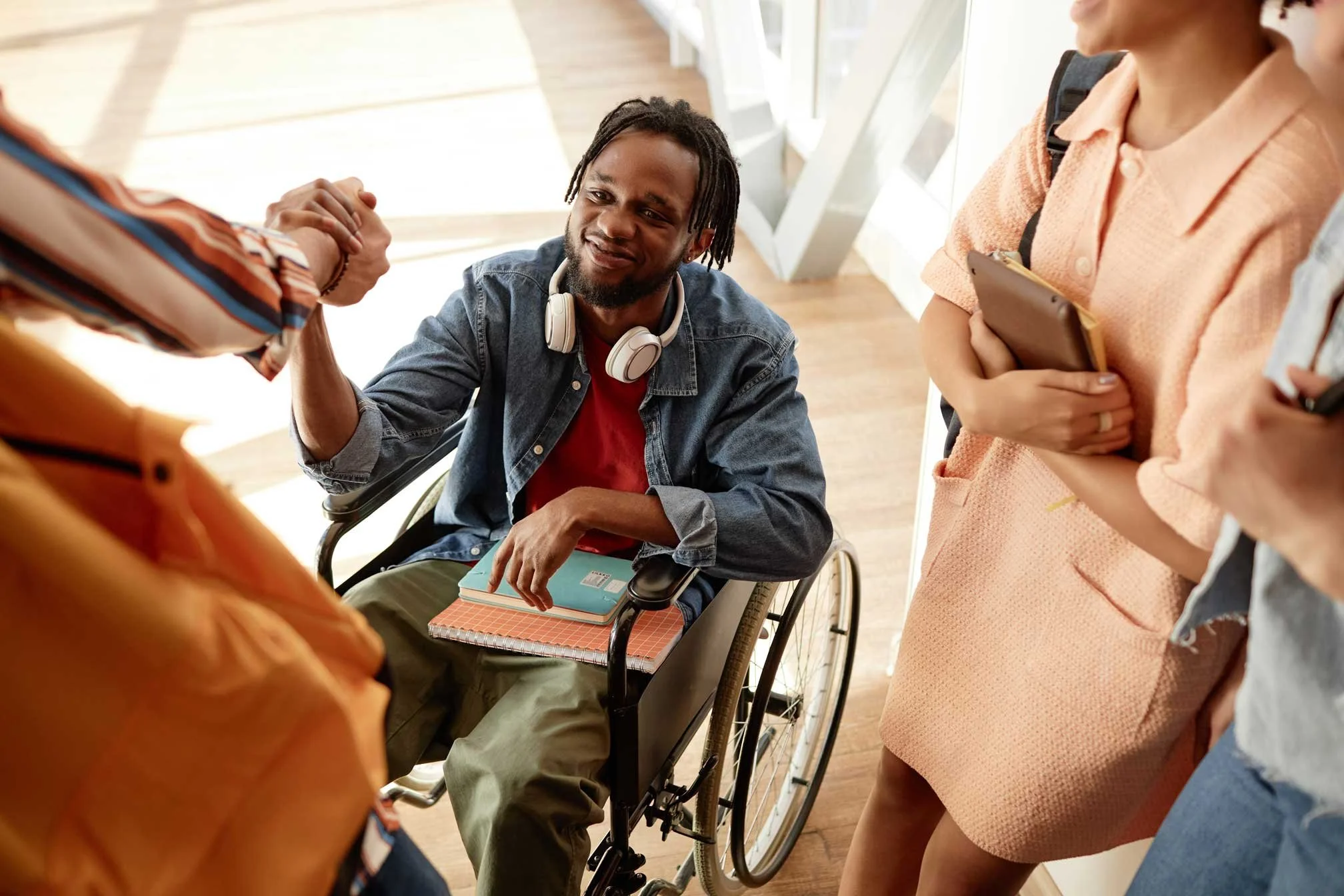 A young black man in a wheel chair with headphone around his next and notebooks in his lap is smiling and shaking hands with his peer | NADSN
