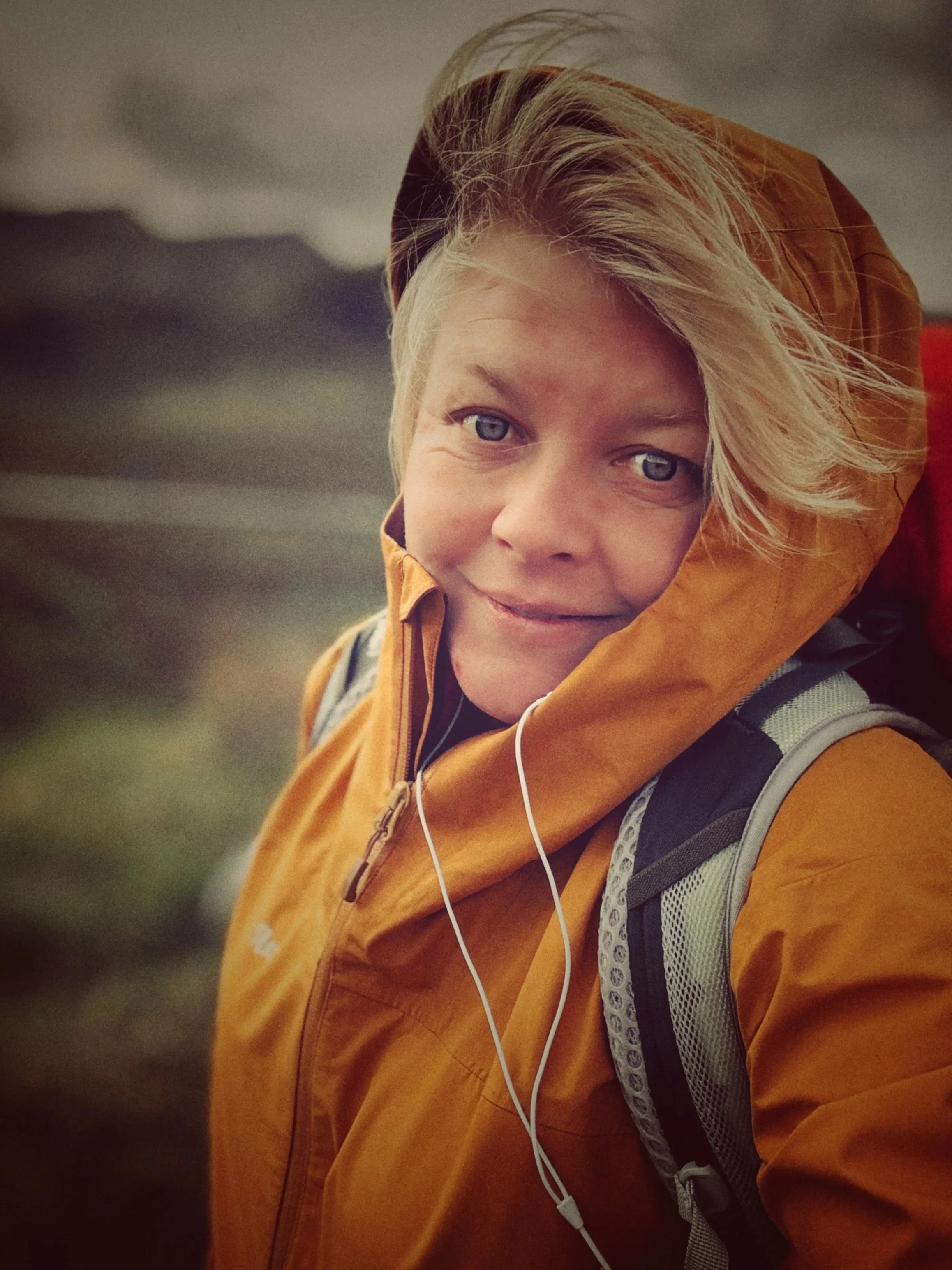A person with blond hair wearing a mustard-colored jacket and a backpack, with earbuds in, outdoors in a natural setting.