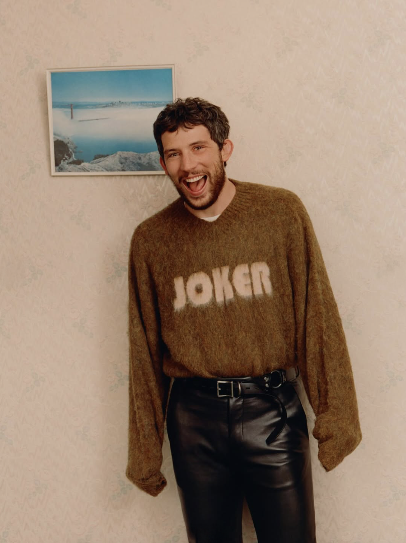 A young man with dark hair and a beard, wearing a brown sweater with the word 'JOKER' and black leather pants, standing against a plain beige wall with a framed beach landscape photograph.