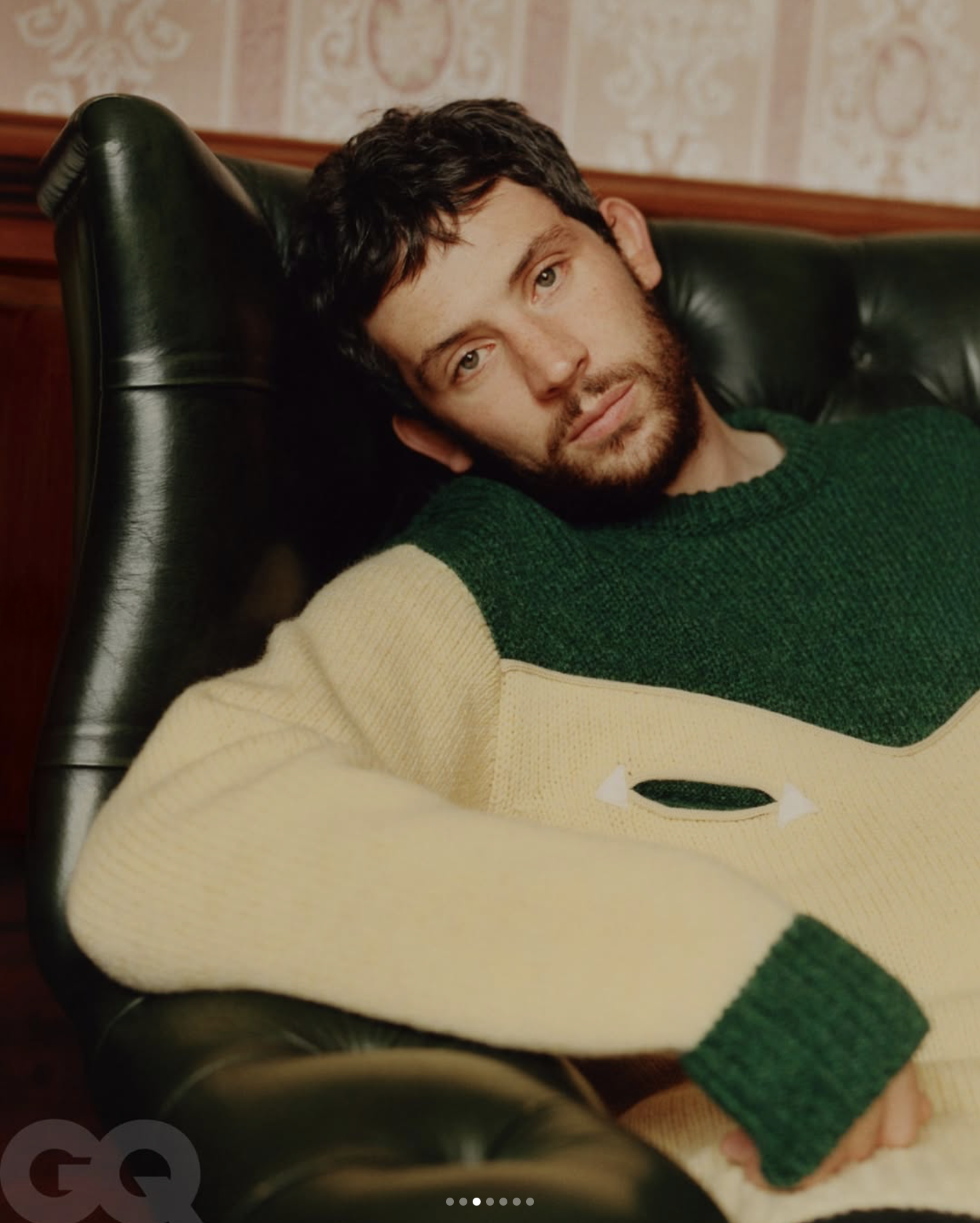 A young man with dark, curly hair and a beard lying on a black leather couch, wearing a cream and green sweater, in a room with ornate wallpaper.