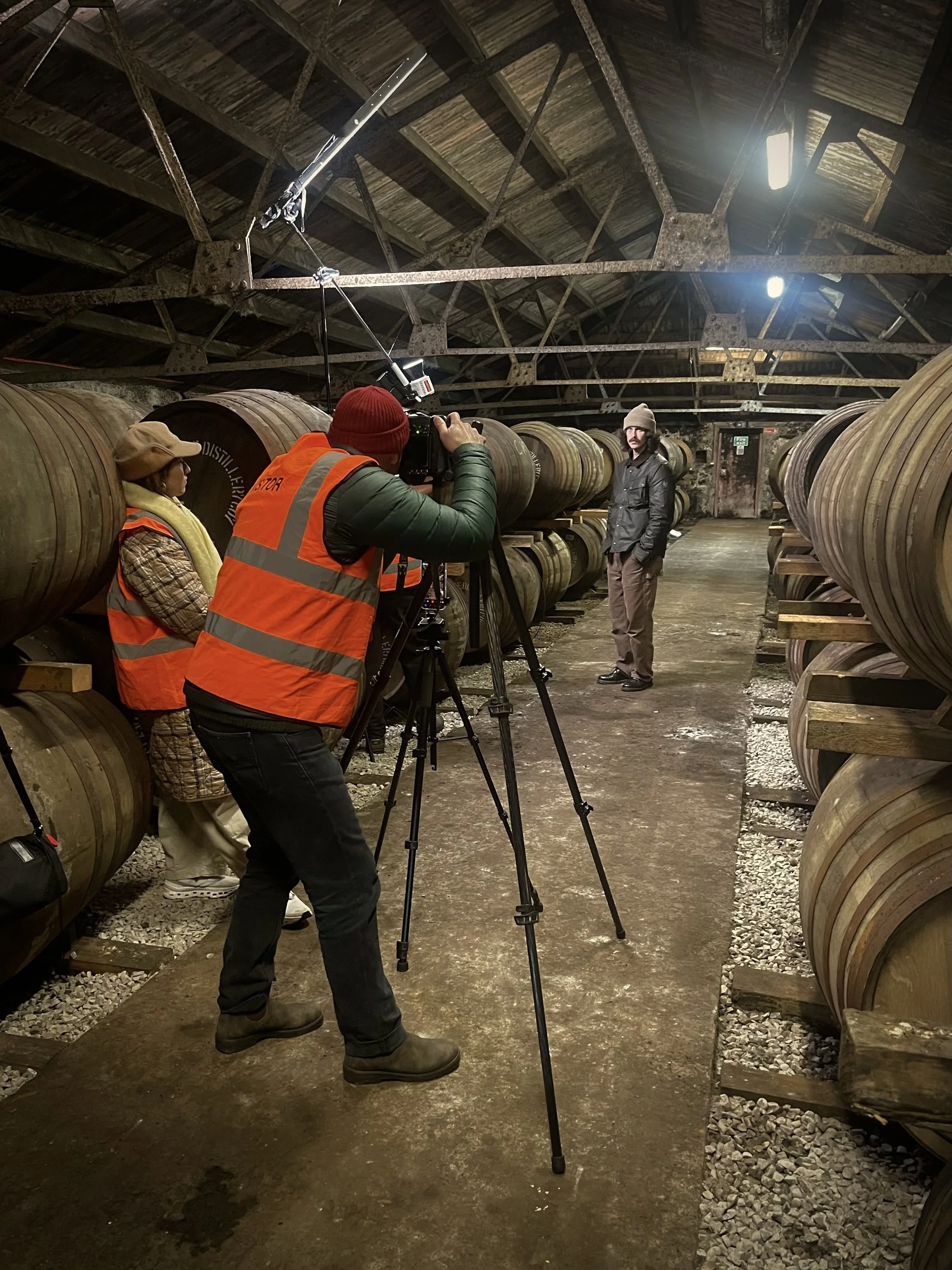 A person being filmed inside a wine cellar with wooden barrels, some individuals are recording and others are posing for the camera.