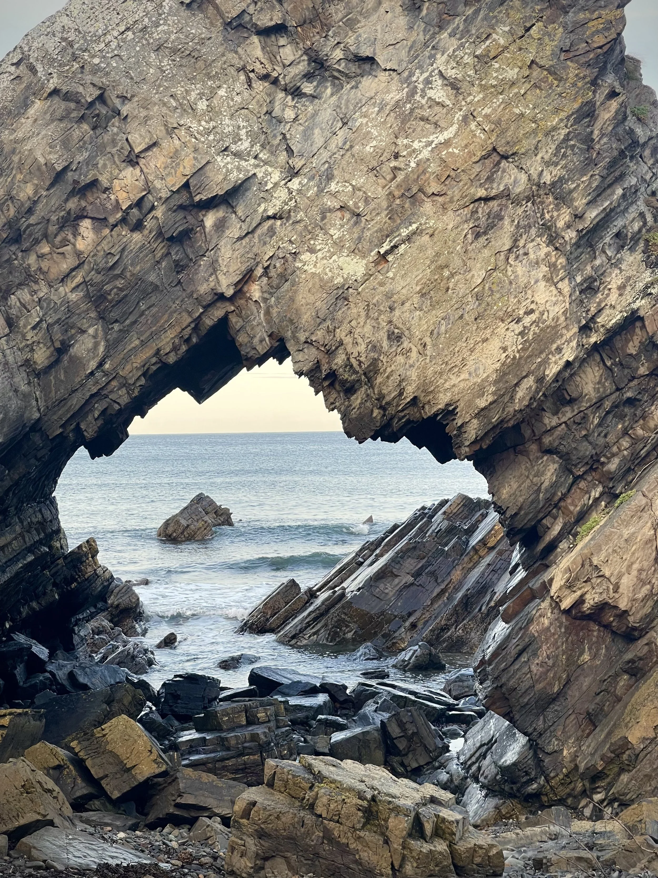 Large rocky arch formation at a beach seen from beneath, with the ocean and a clear sky in the background.