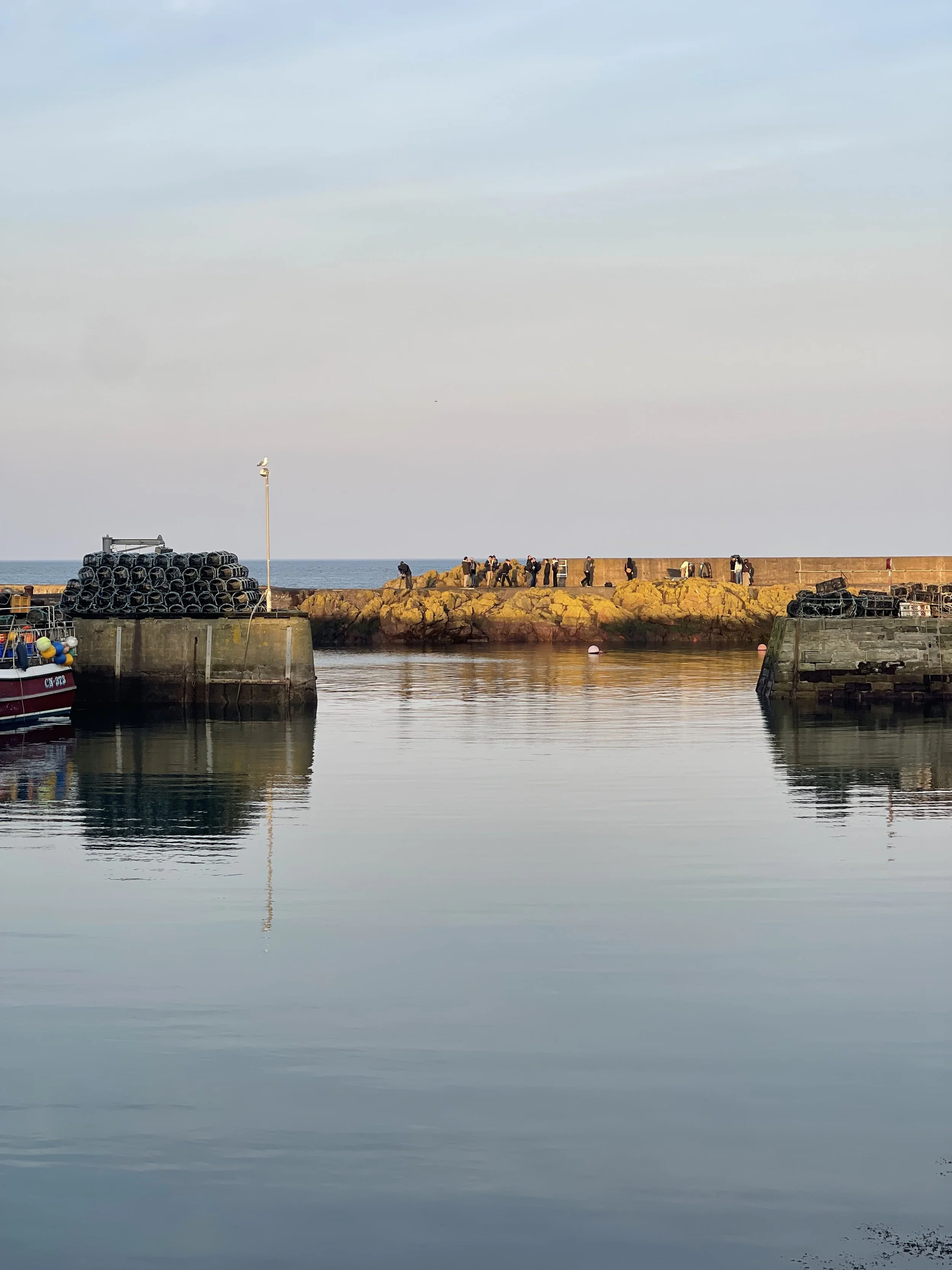 A harbor scene with calm water, rocks along a pier, and several people walking on a breakwater. Boats are docked on the left, and the sky is partly cloudy.