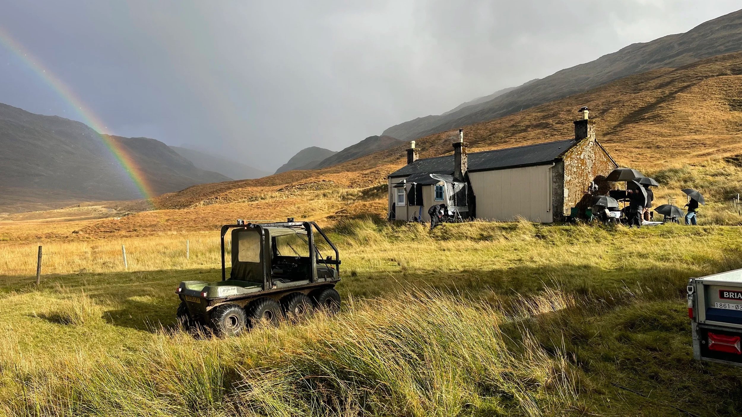 Film crew shooting scenes outside a house in a rural landscape with mountains and a rainbow in the background. Several crew members hold umbrellas, and filming equipment is set up near the house.