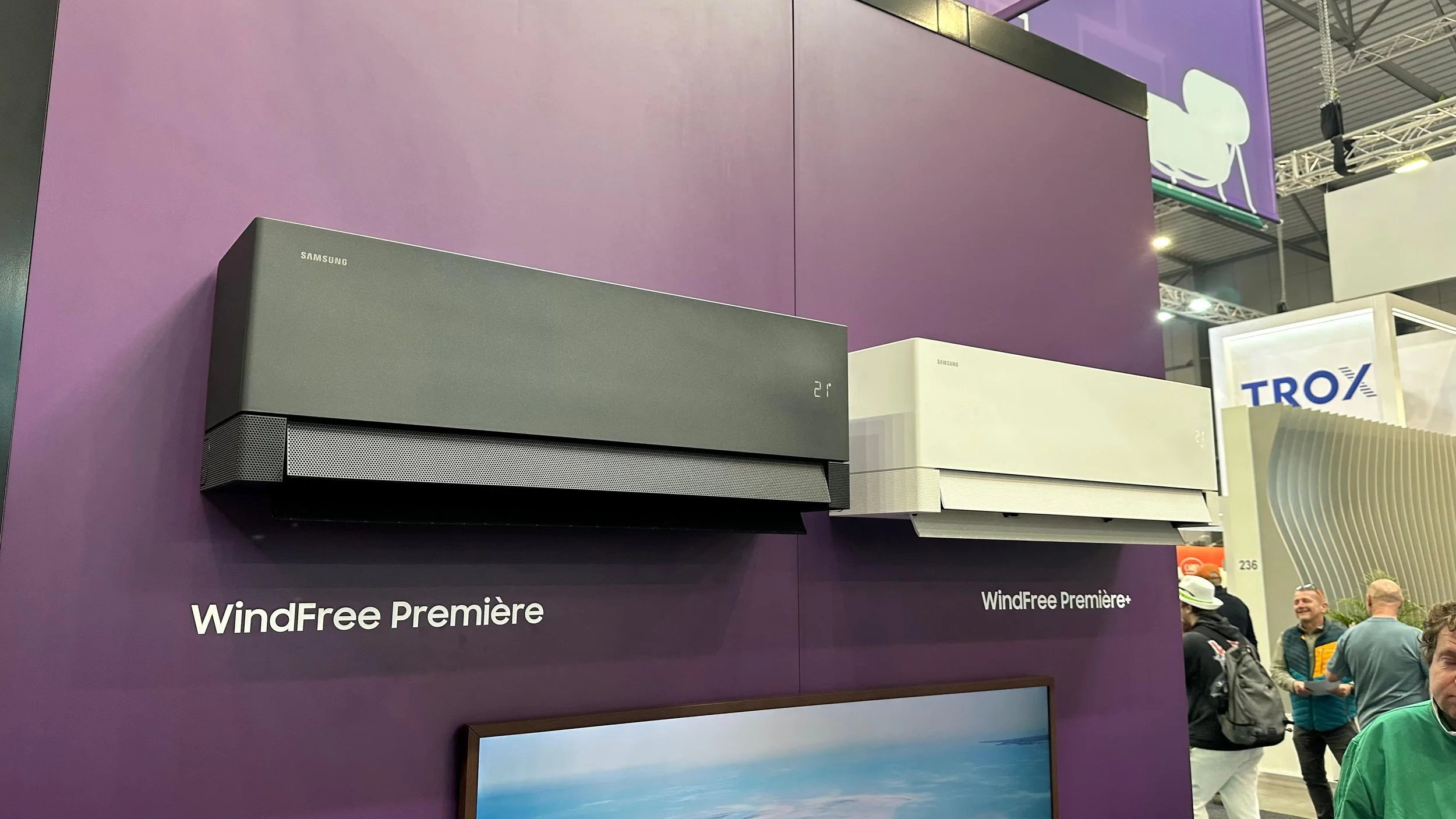 Výstavná prezentácia klimatizácií Samsung WindFree Première, kde sú dve jednotky namontované na fialovej stene, v pozadí sa nachádza skupina ľudí; scéna zachytáva veľtržnú expozíciu moderných HVAC riešení.