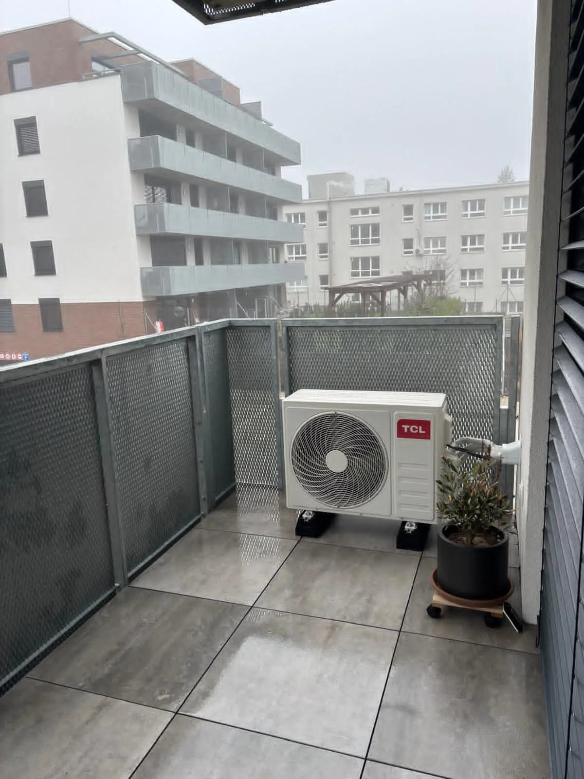 Malý balkón s mokrou dlažbou, kovovým zábradlím, klimatizačnou jednotkou TCL a črepníkovou rastlinou na pojazdnom stojane s viditeľnými kolieskami, s výhľadom na moderné bytové domy počas daždivého dňa, vhodný pre rezidenčné použitie klimatizácie.